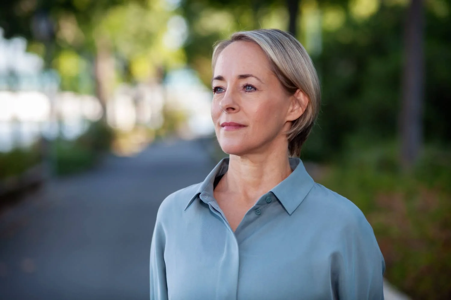 Julie Morrow standing outdoors on a tree-lined path, wearing a light blue button-up shirt, looking contemplative.