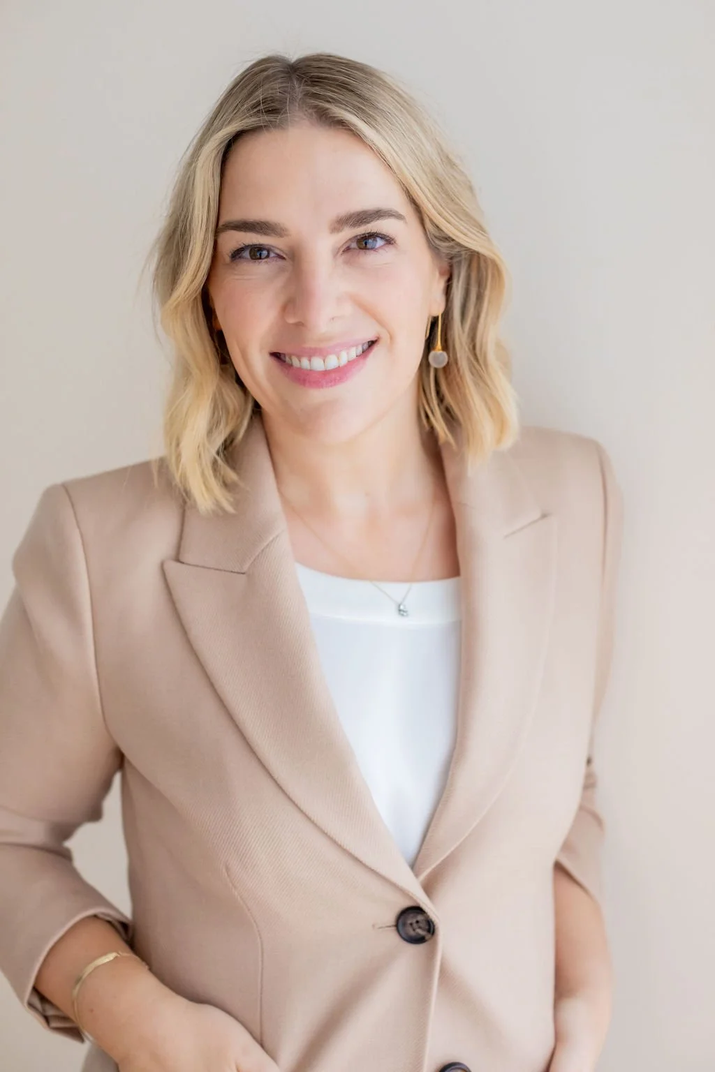 A smiling woman with shoulder-length blonde hair, wearing a beige blazer over a white top, with a necklace and an earring, standing against a plain light-colored background.