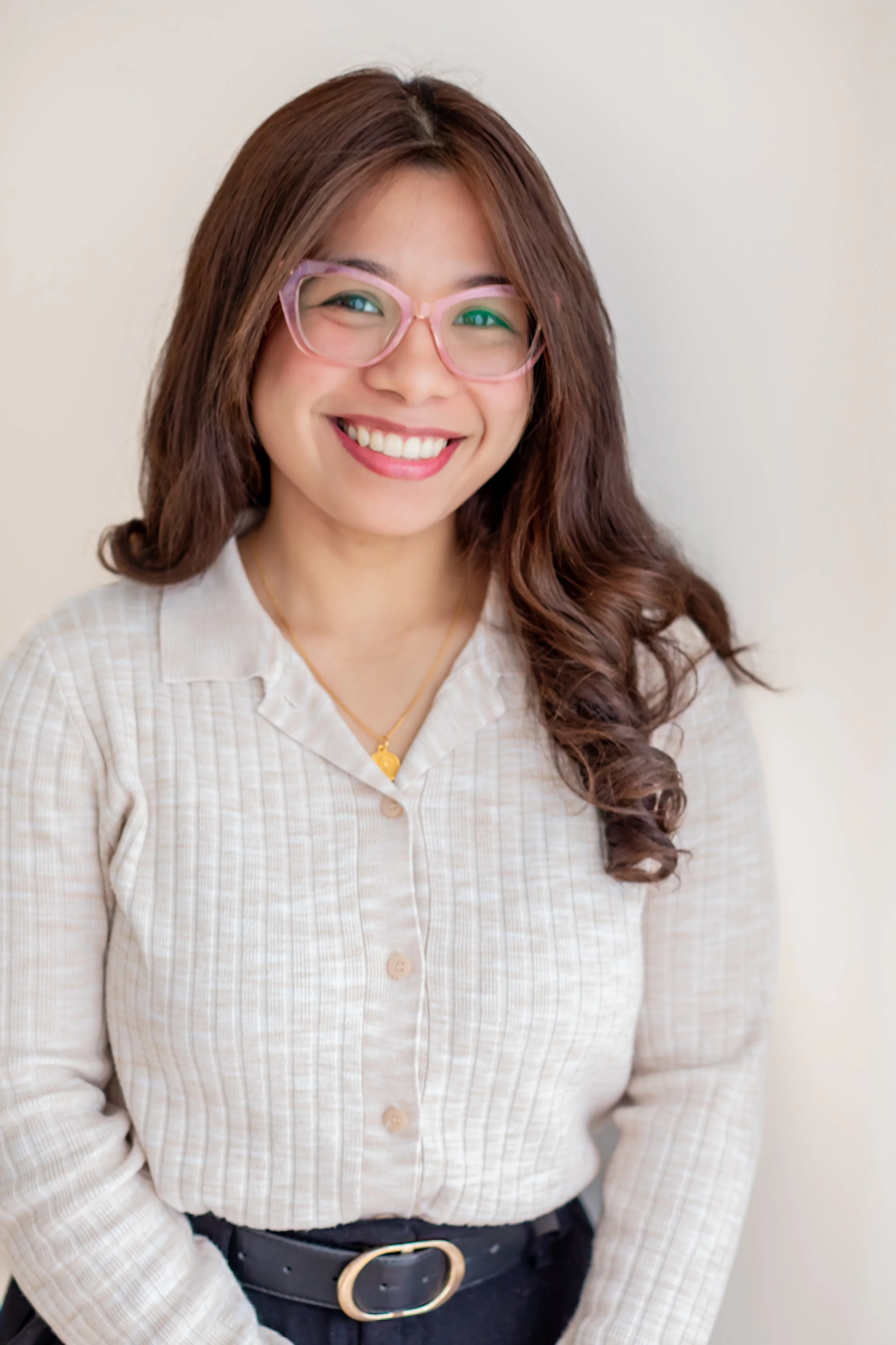 A woman with long, wavy brown hair wearing light pink glasses and a cream-colored, checkered blouse with a gold necklace. She is smiling and standing against a plain background.