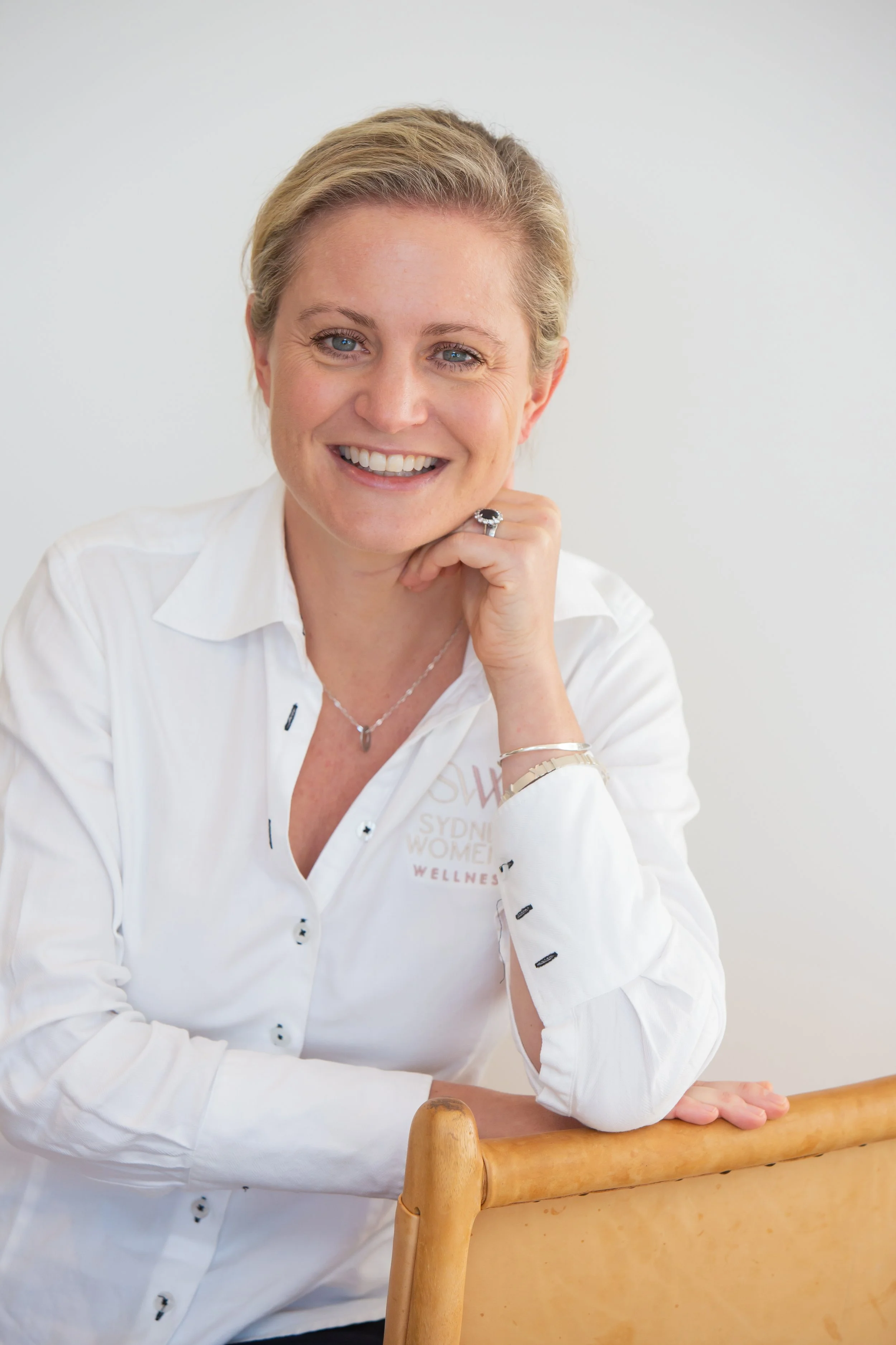 Smiling woman with short blonde hair, wearing a white button-up shirt with partial text on the chest, sitting with her left hand resting on her chin, jewelry on her fingers and wrist, in a room with plain white background.