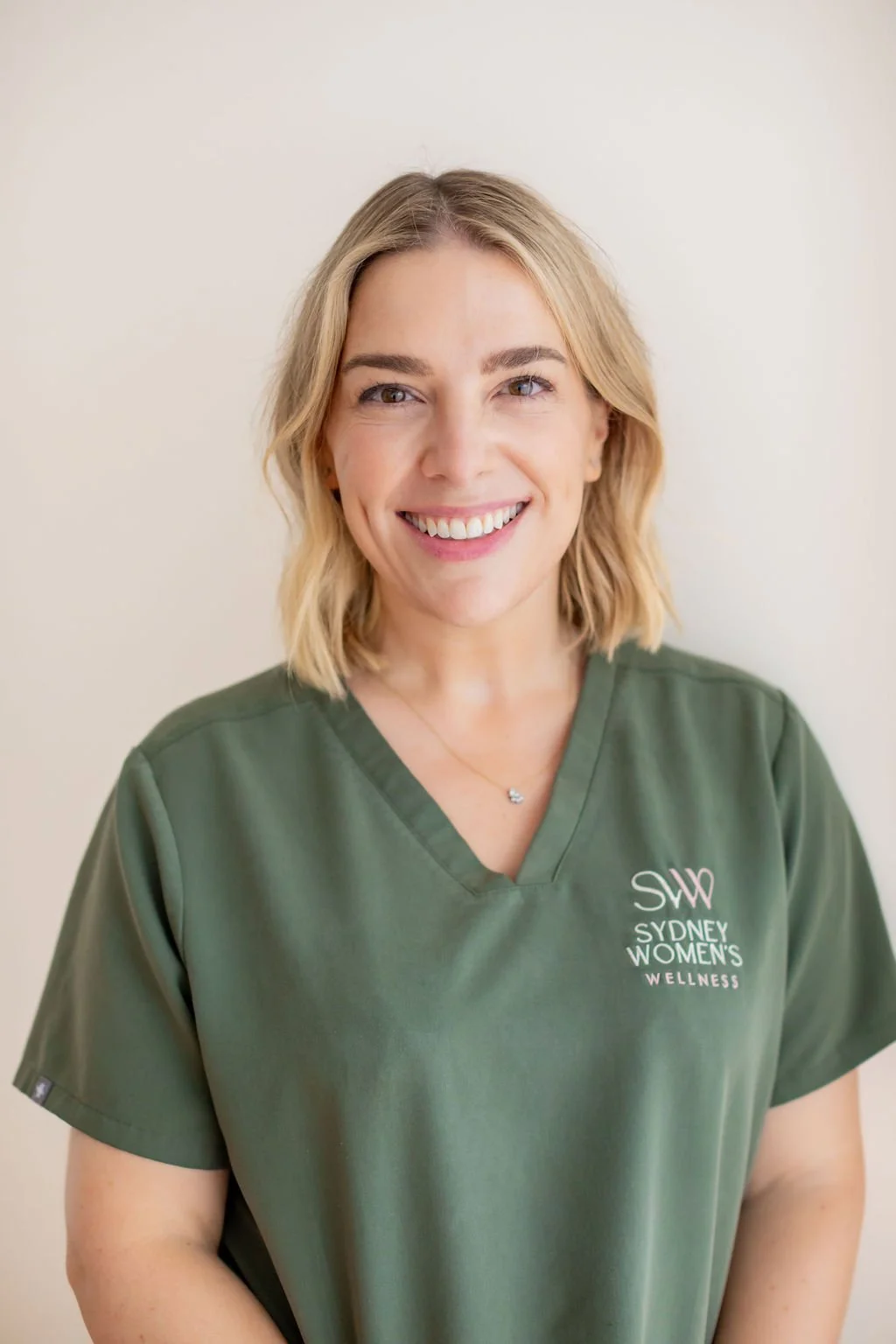 A smiling woman with blonde hair wearing a green medical scrub top with 'Sydney Women's Wellness' embroidered on it.