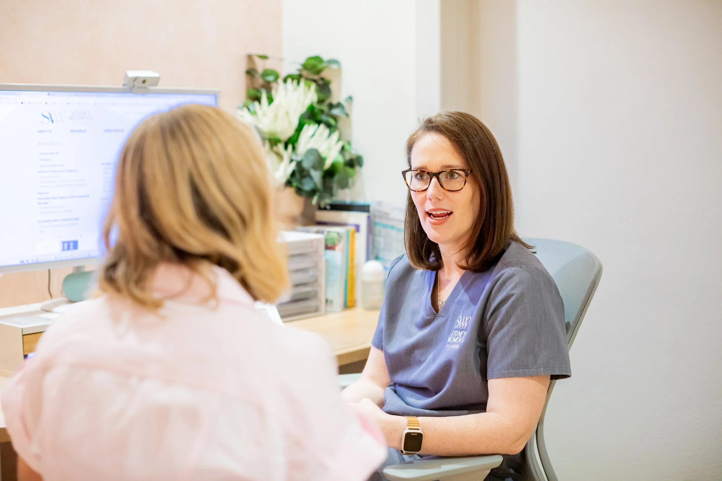 A woman in medical scrubs sitting at a desk and talking to a patient with blonde hair. The woman is wearing glasses and has shoulder-length brown hair. There are medical books and a monitor on the desk, with some flowers in the background.