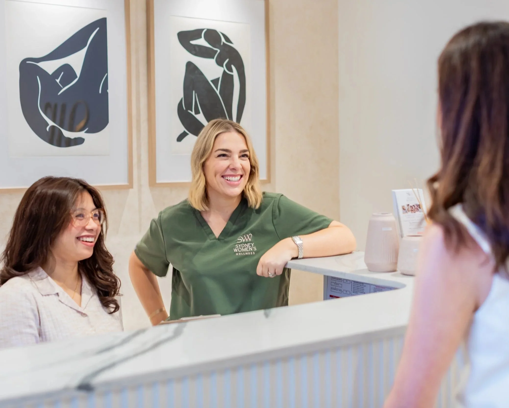 Two women, one weaing a white blose and the other wearing a green shirt leaning on a reception desk, speaking to another woman with brown hair and a white top.