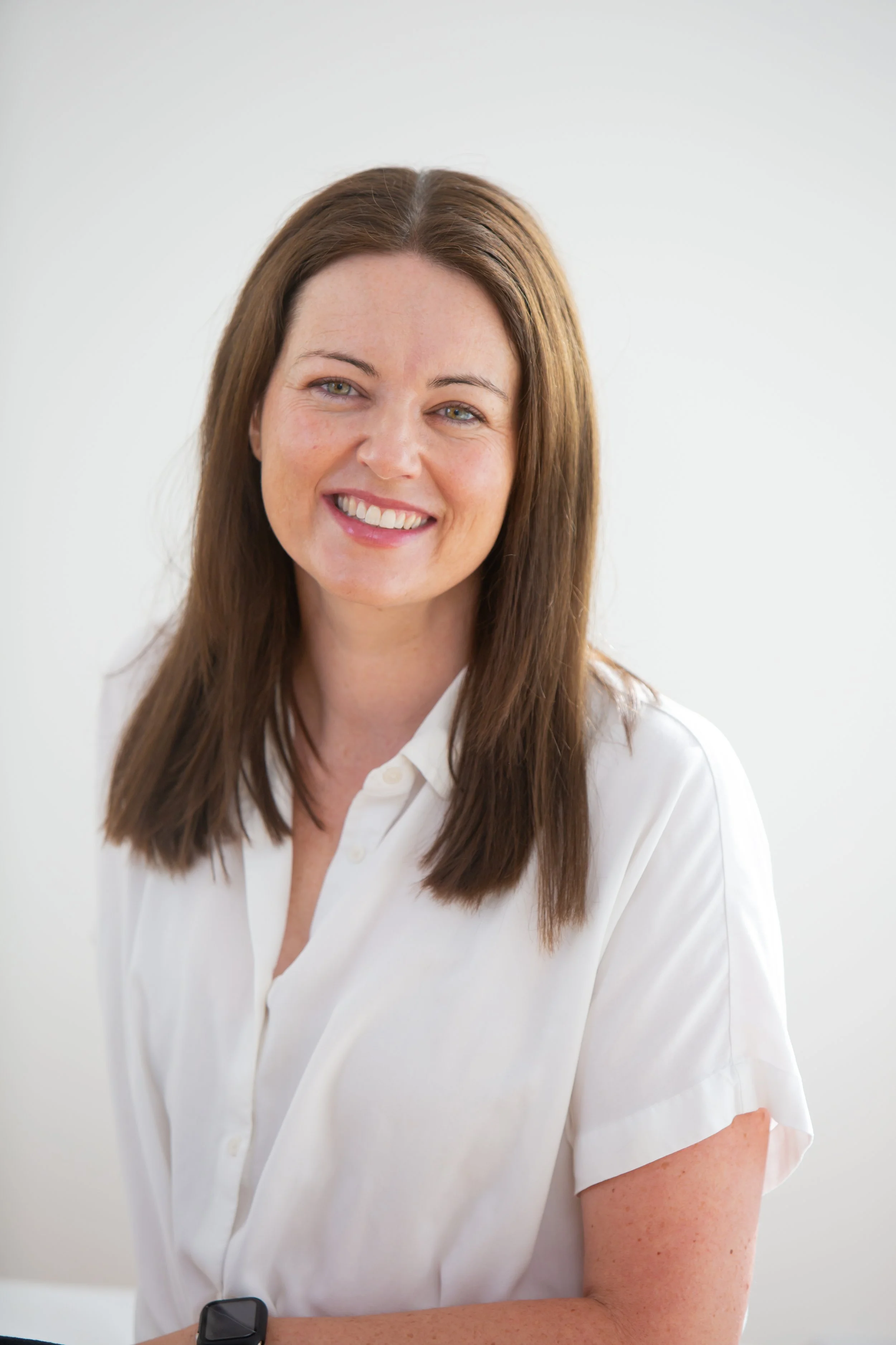 A woman with long brown hair wearing a white shirt and a black smartwatch, smiling while standing against a plain white background.