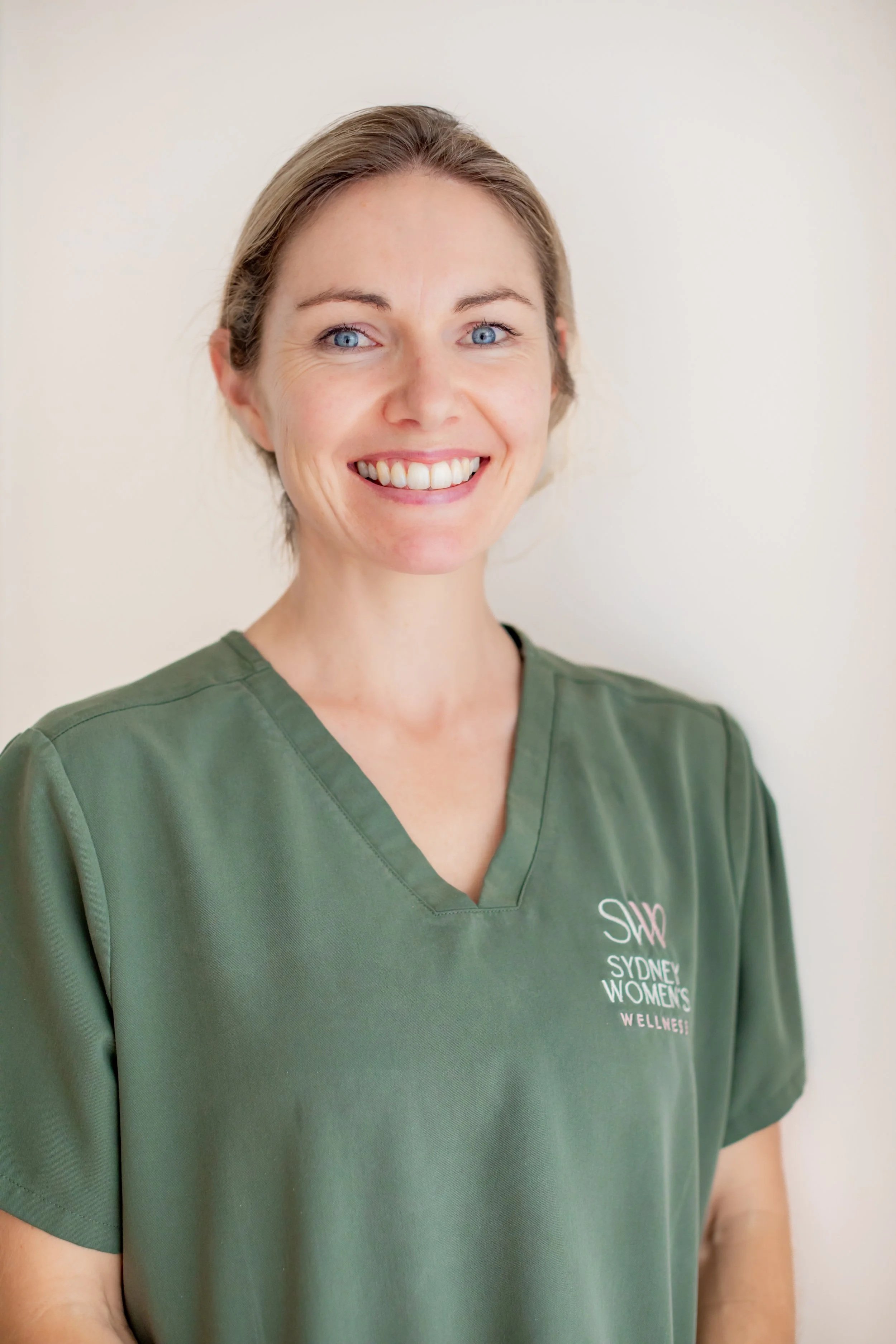 A smiling woman with short light brown hair, wearing green medical scrubs with a logo that reads 'Sydney Women's Wellness' on the chest, standing against a plain white wall.