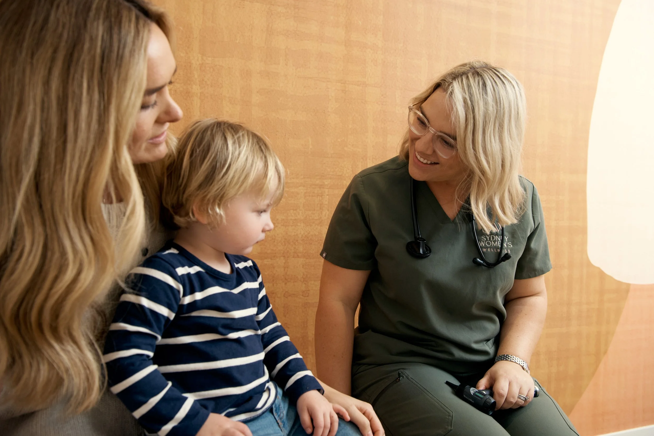 A female medical professional in green scrubs, wearing glasses and a stethoscope, is smiling and talking with a woman and a young boy. The woman has long, wavy, reddish-blonde hair, and the boy has blonde hair and is wearing a striped navy and white shirt. They are sitting on a bench in a medical or healthcare setting with a wood-patterned wall in the background.