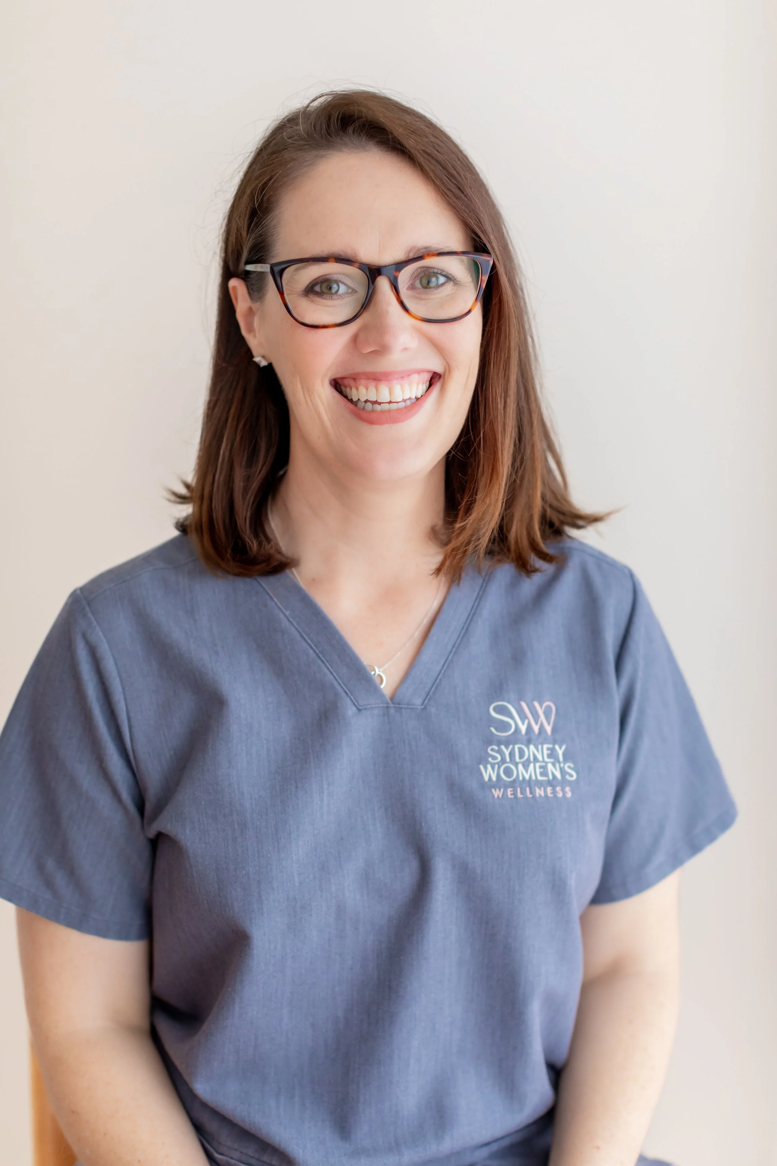A woman with brown hair, glasses, and a big smile, wearing a blue scrub top with the logo 'Sydney Women's Wellness'