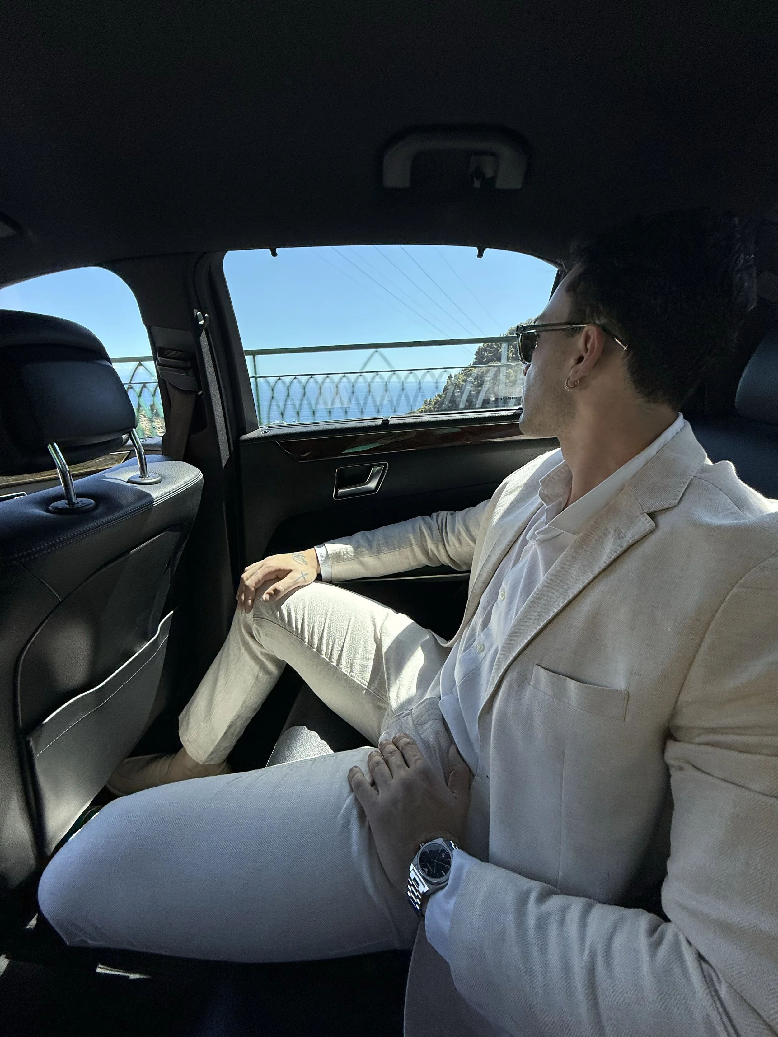 A man sitting in the backseat of a car, looking out the window at a bridge with a scenic ocean view.