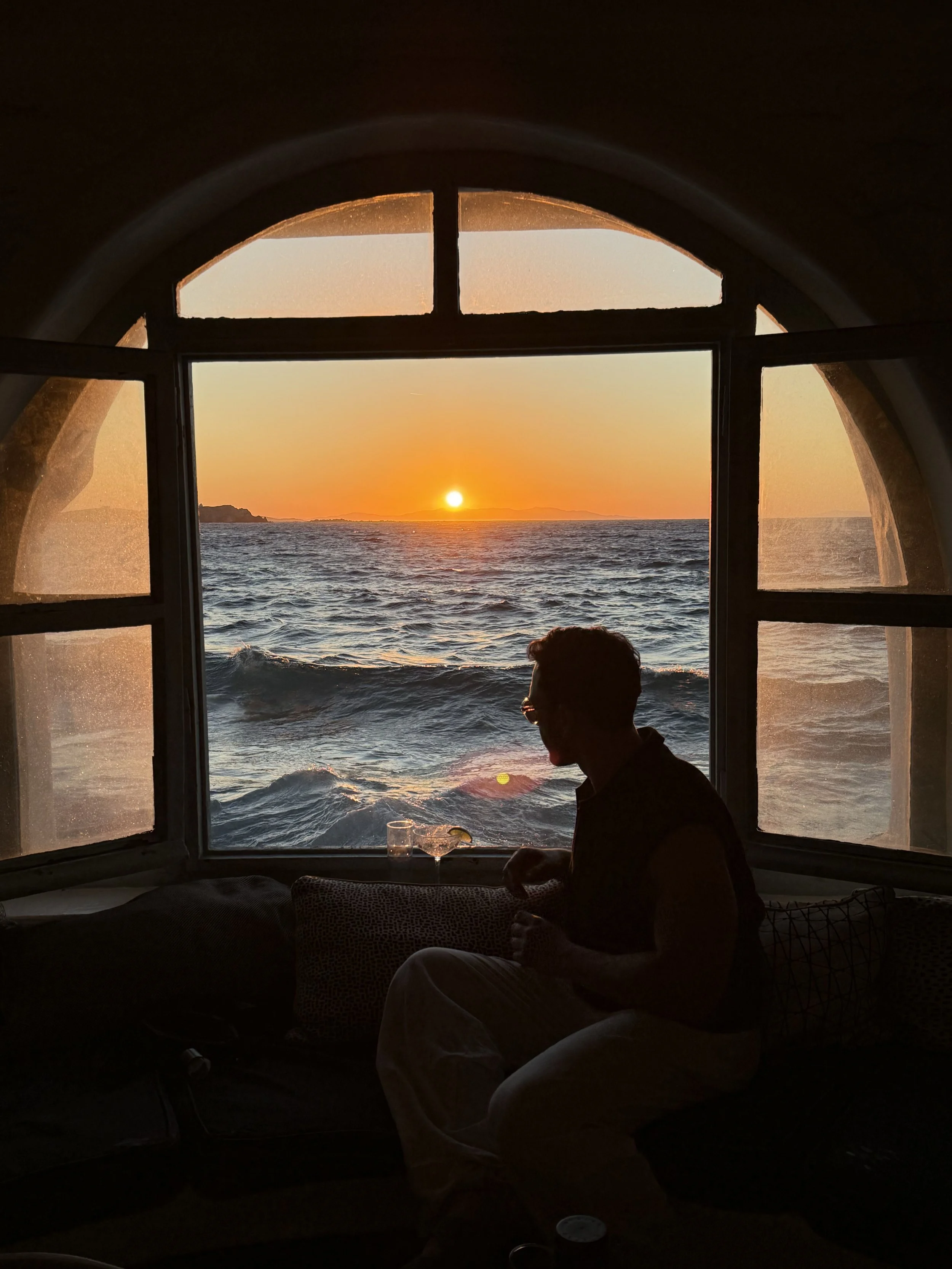 Silhouette of a person sitting inside a room, looking out a window at the sunset over the ocean with waves and distant land.