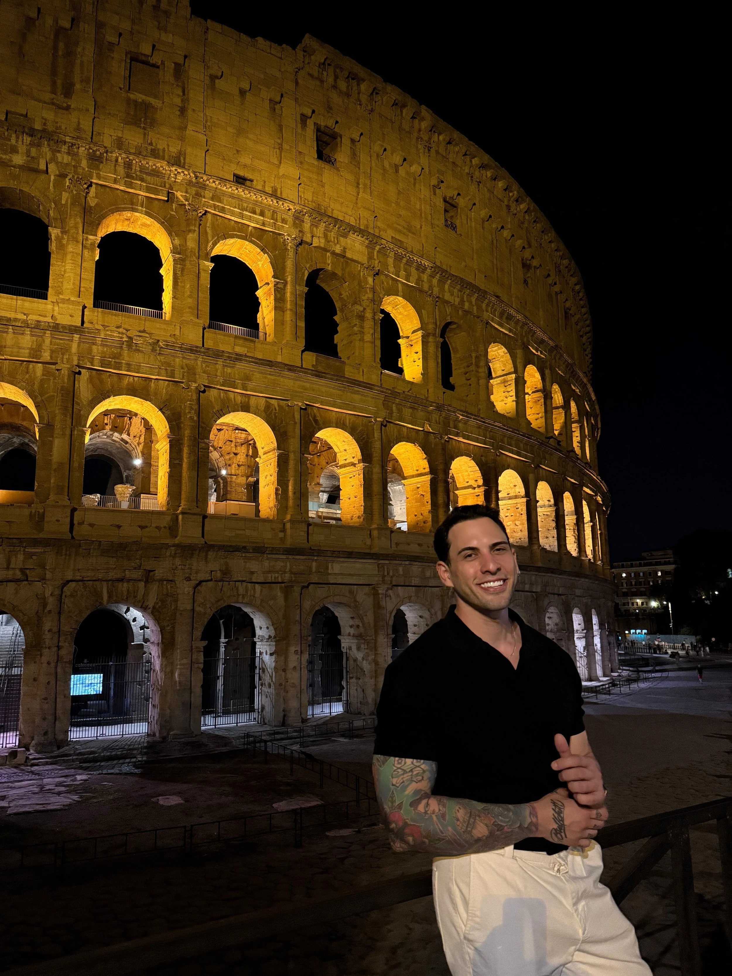 A smiling man with tattoos on his arms, wearing a black shirt and light-colored pants, standing in front of the illuminated Roman Colosseum at night.