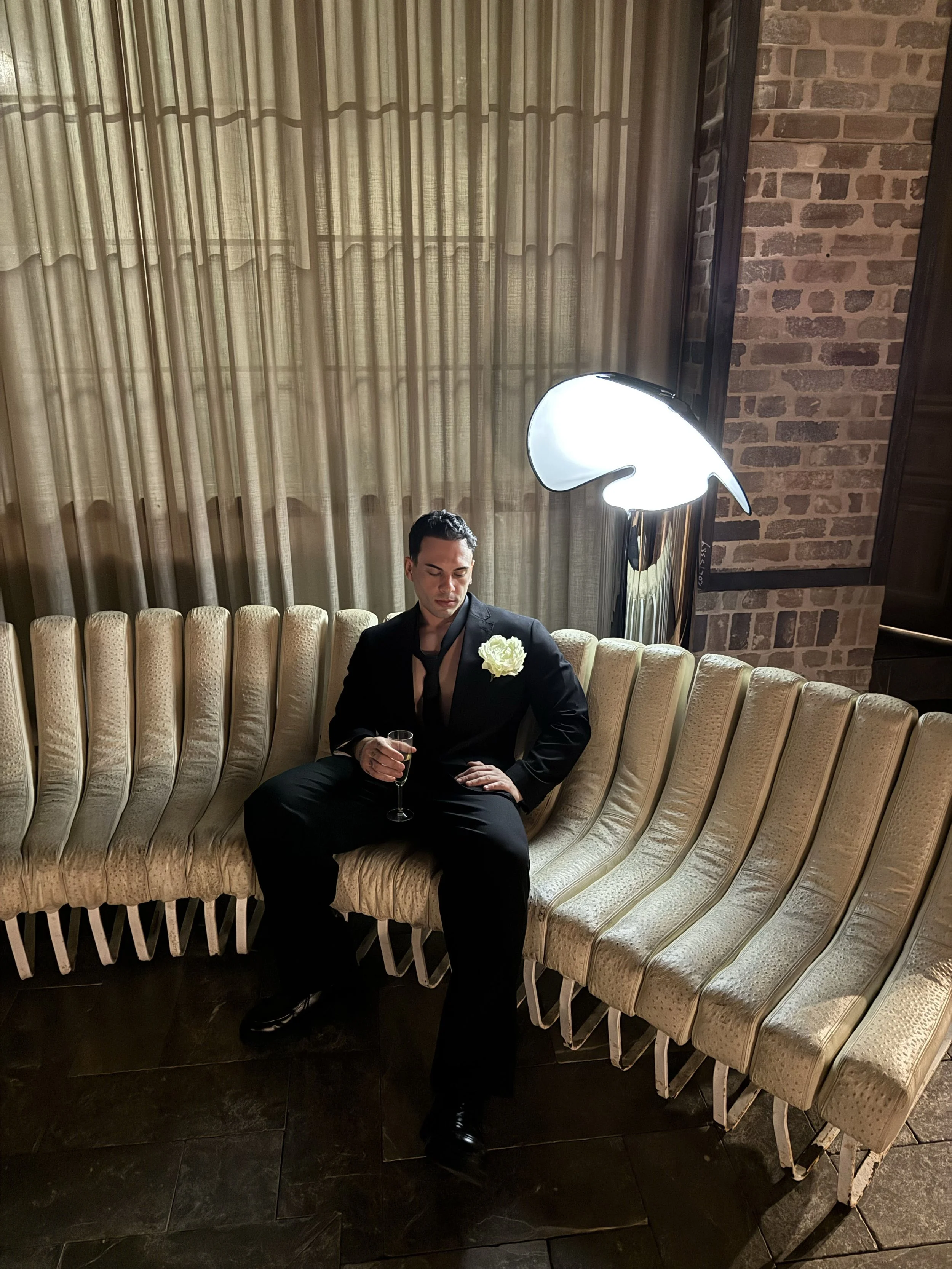 A man in a black suit with a white flower on his lapel, sitting on a beige curved sofa in a dimly lit room. He is holding a glass of champagne in his right hand, with one leg crossed over the other. There is a large, modern, bird-shaped floor lamp overhead, casting light onto him. The background features beige curtains and a brick wall.