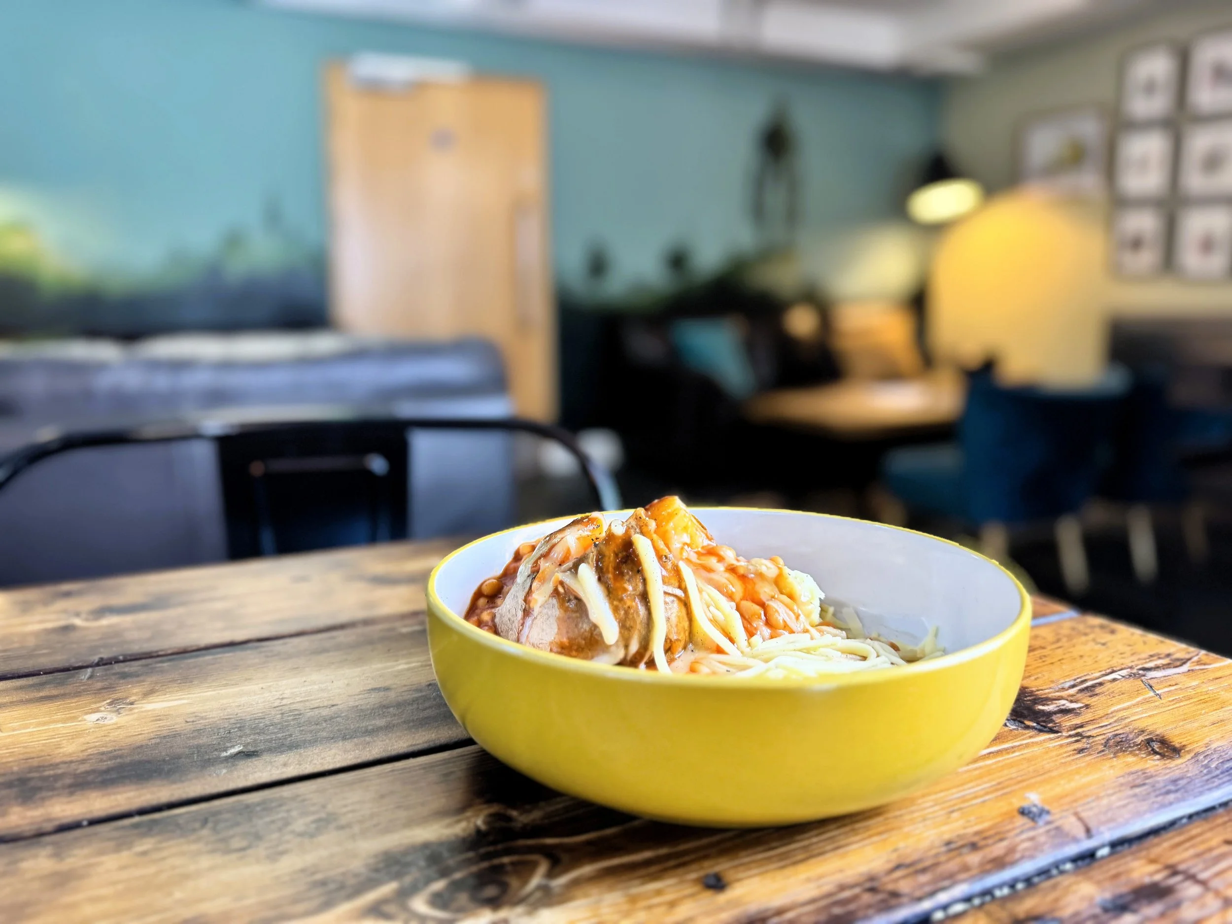 A jacket potato with cheese on a wooden table in a cosy, decorated room with a bed, seating area, and framed pictures Lucy's Little Bakehouse, Dean Clough, Halifax.