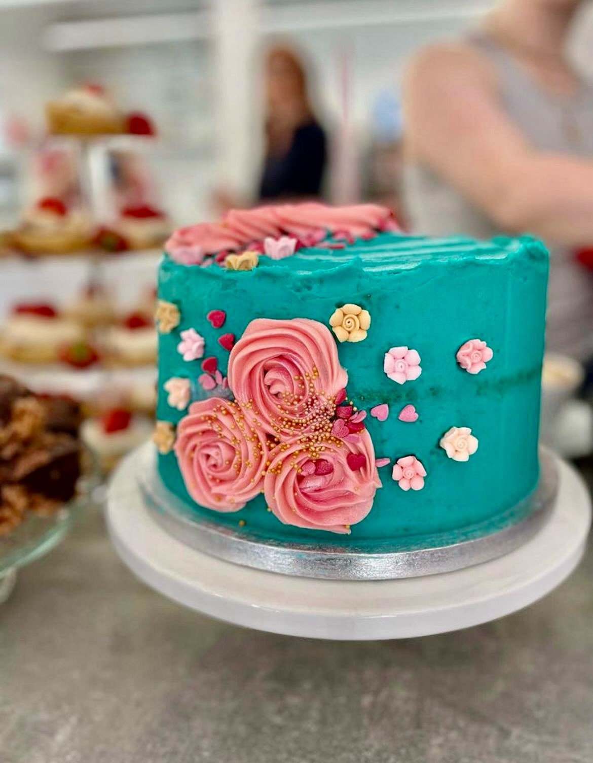 A colorful birthday cake with pink roses and small pink and white flowers on a silver cake board, in Lucy's Little Bakehouse, Dean Clough, Halifax.