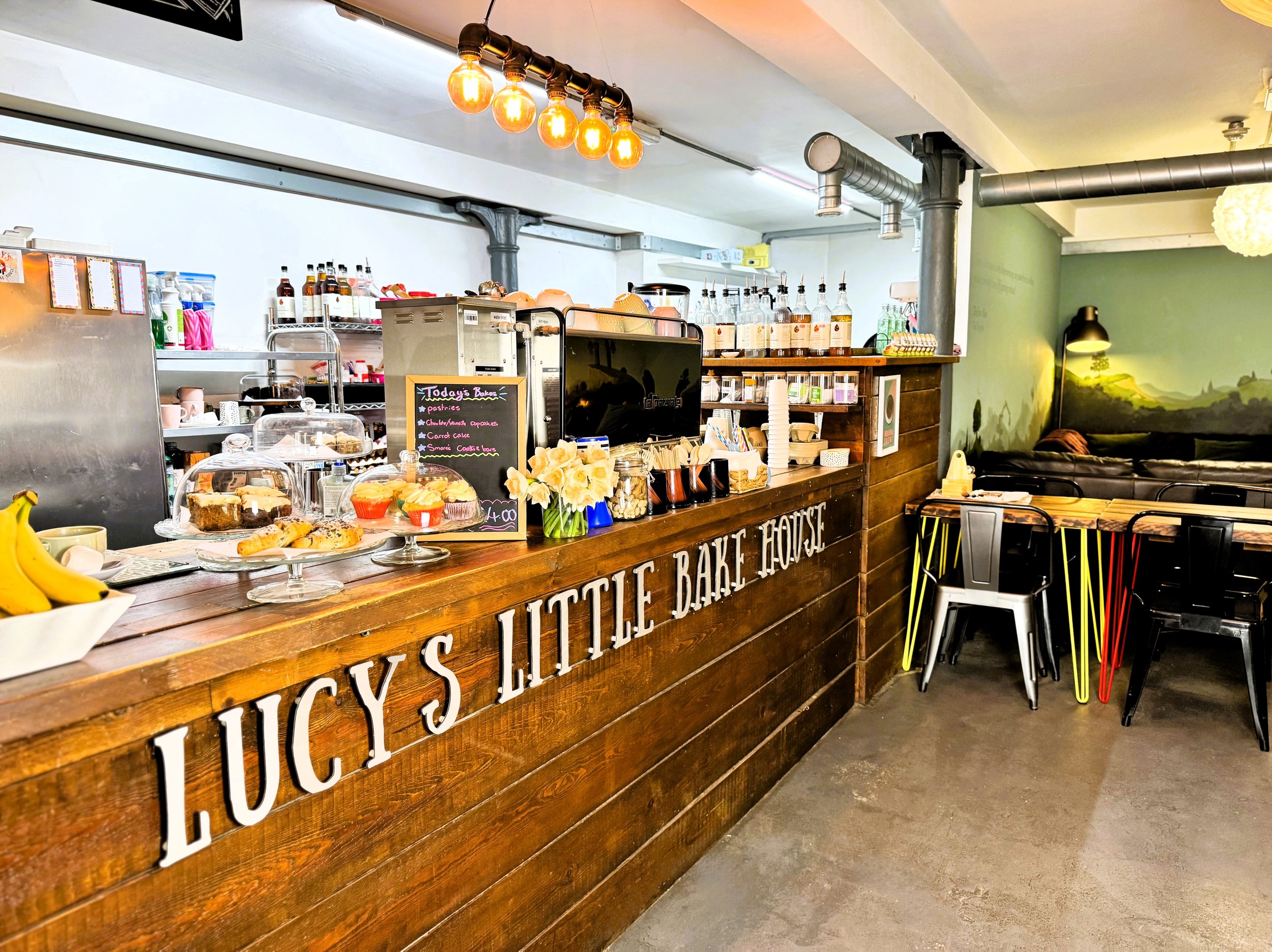 Interior of a coy bakery with a wooden counter that reads 'Lucy's Little Bake House,' displaying baked goods like pastries and cupcakes, with a chalkboard menu, flowers, and coffee supplies. In the background, shelves with bottles and baking ingredients, and a small dining area with tables and chairs.