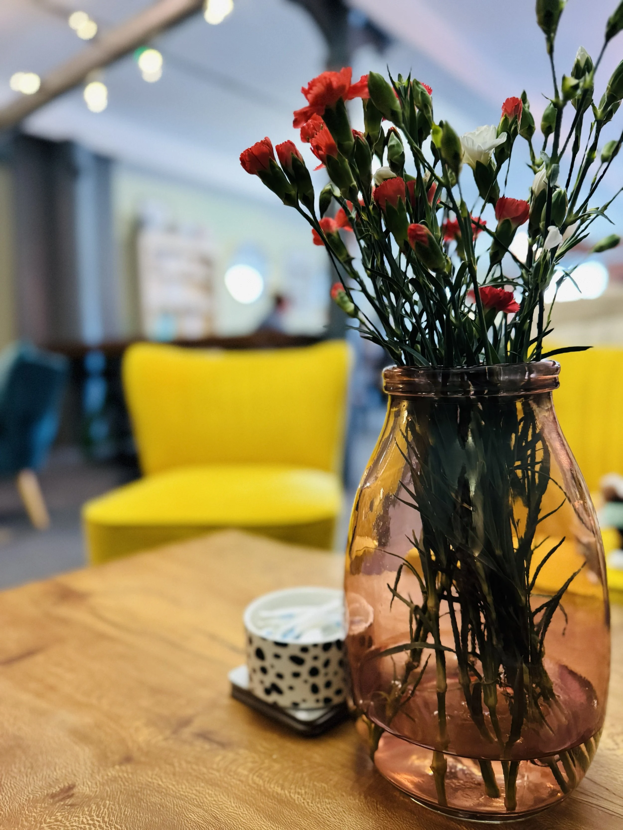 A pink glass vase with a bouquet of red and white flowers sits on a wooden table in a cozy, modern interior space. The background includes blurred yellow and teal chairs and ambient lighting.