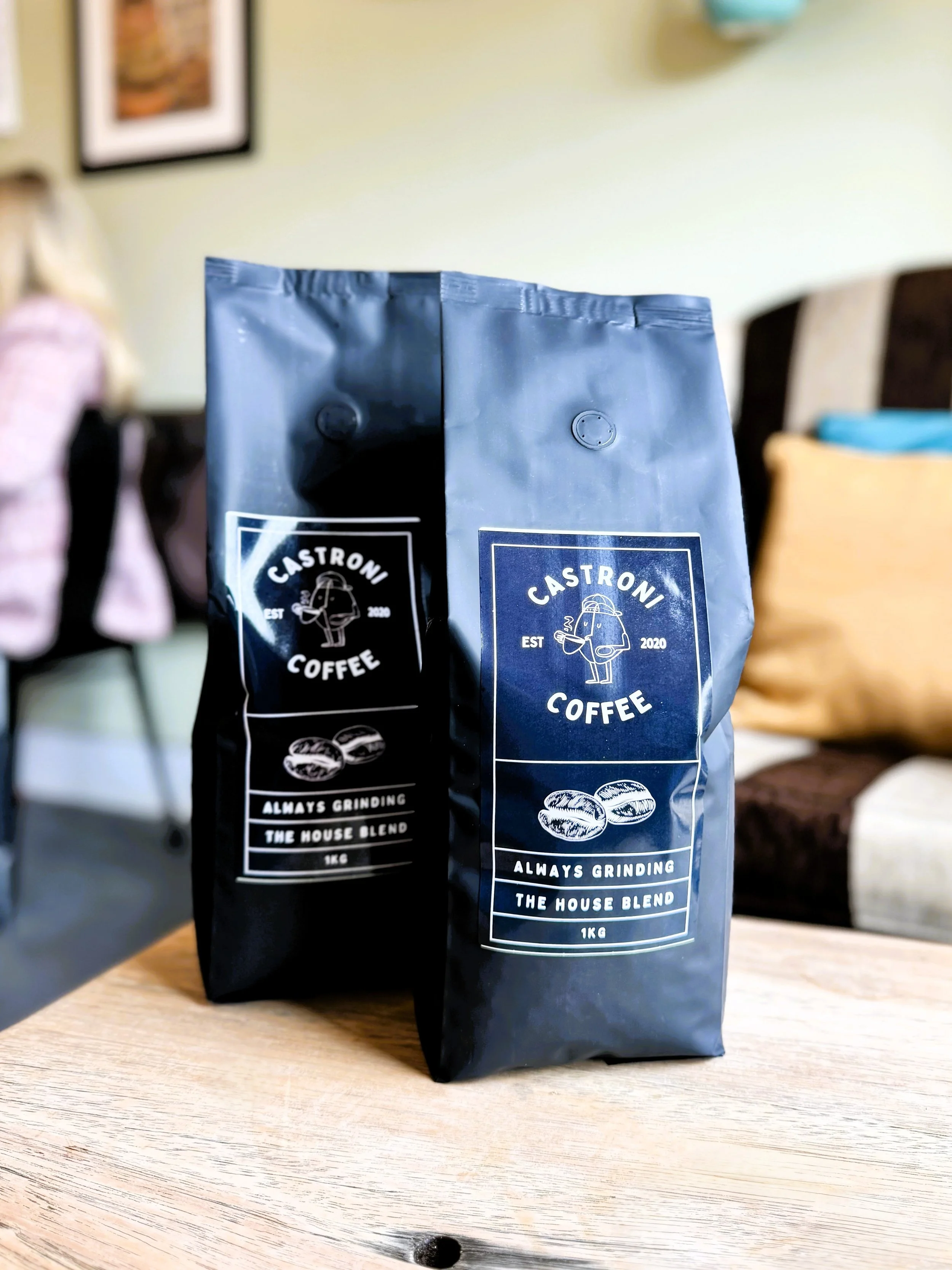 Two bags of Castroni Coffee with a logo of a coffee mug and a book, labeled 'Always Grinding, The House Blend, 1kg,' placed on a wooden table with a blurred background of a cosy indoor setting in Lucy's Little Bakehouse Halifax