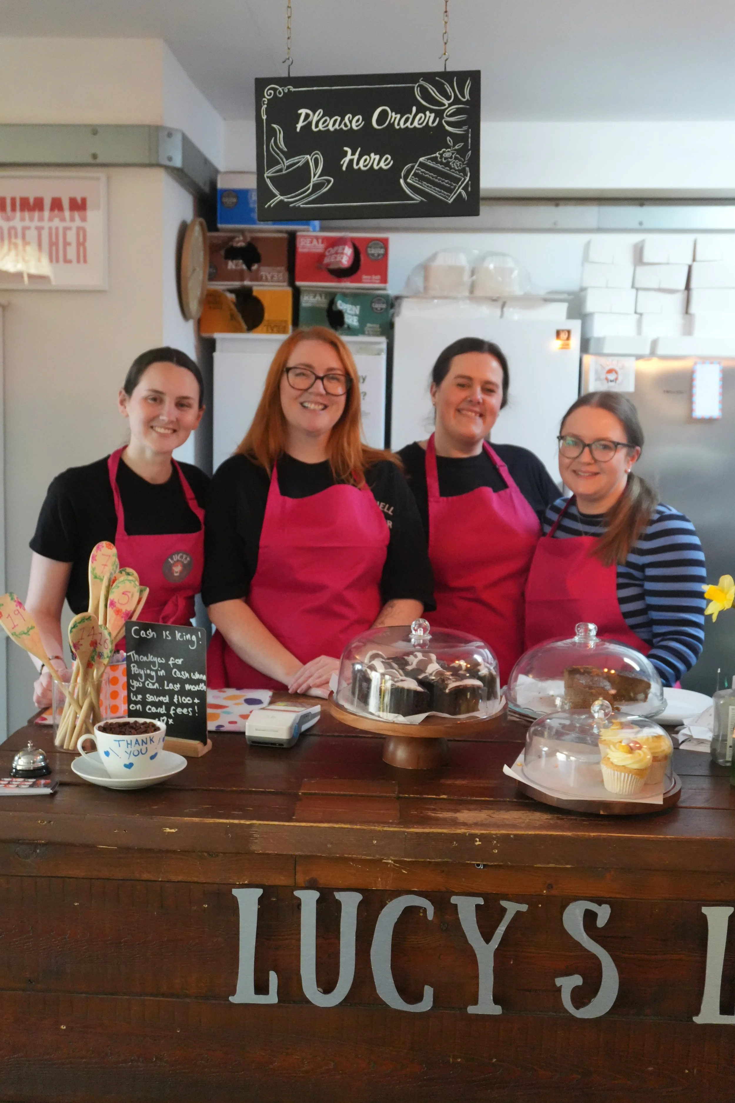 Four women behind a bakery counter with cakes and cupcakes, wearing pink aprons, in Lucy's Little Bakehouse, Dean Clough, Halifax