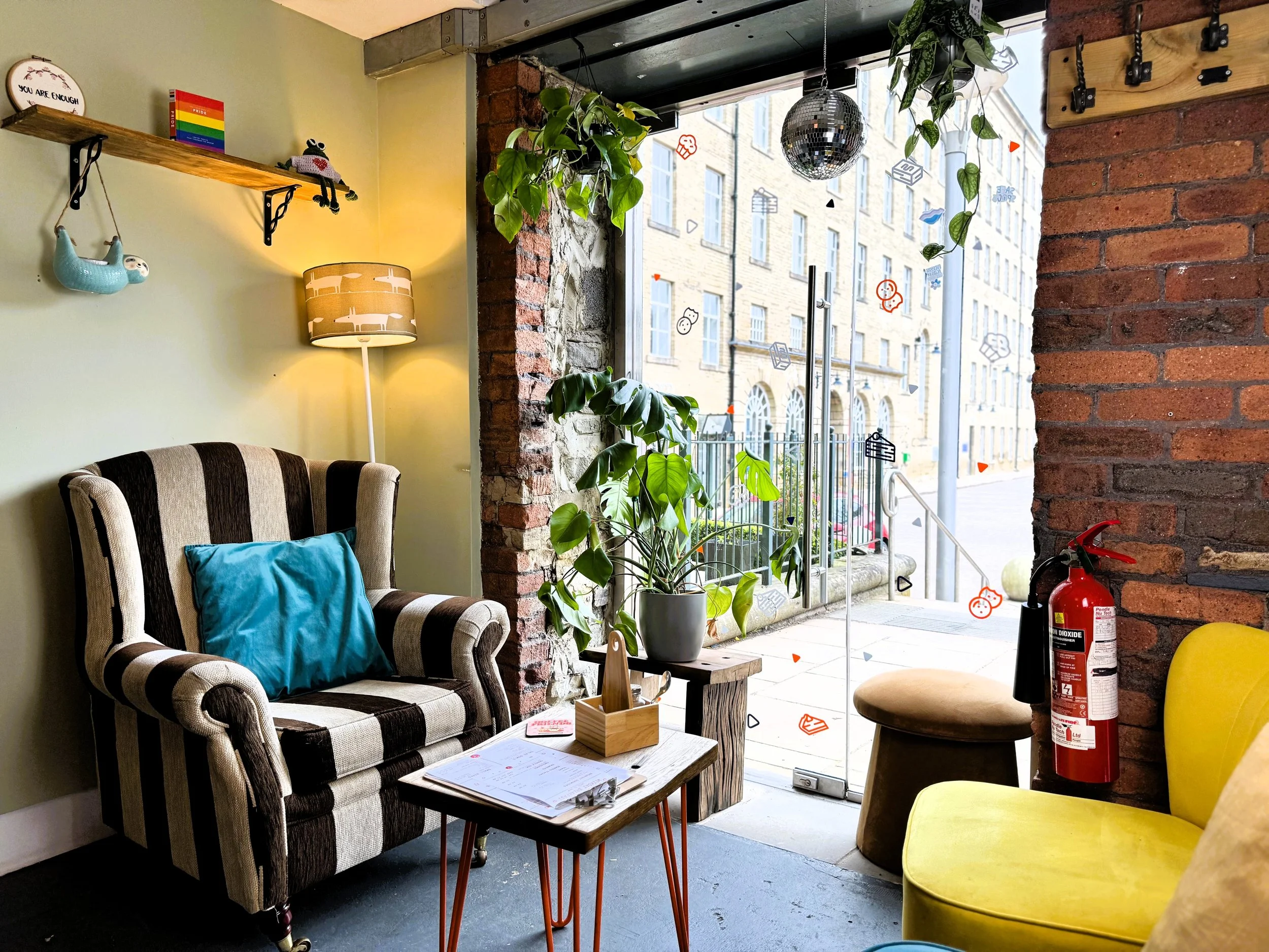 Cosy cafe corner with striped armchair, yellow sofa, small coffee table, potted plant, hanging plants, brick walls, large window with city view, decorative items in Lucy's Little Bakehouse, Dean Clough, Halifax.