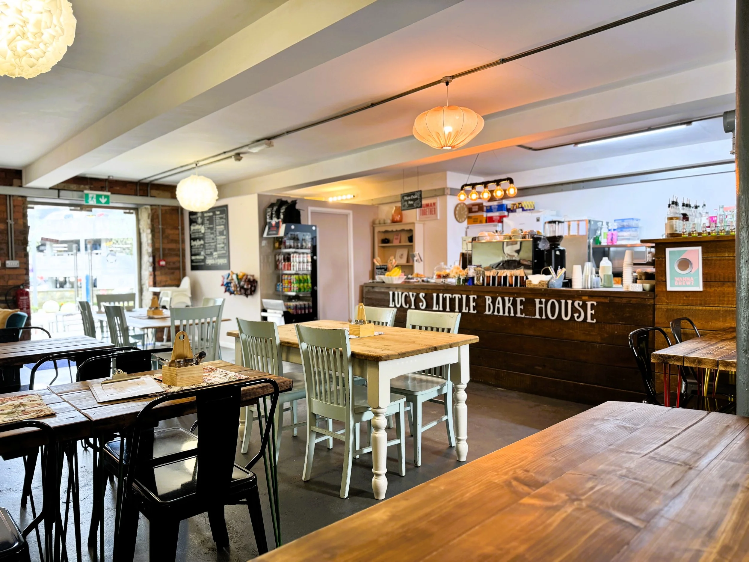 Interior of a cosy cafe in Dean Clough Halifax with wooden tables and chairs, a counter reading "Lucy’s Little Bake House," and warm lighting fixtures.
