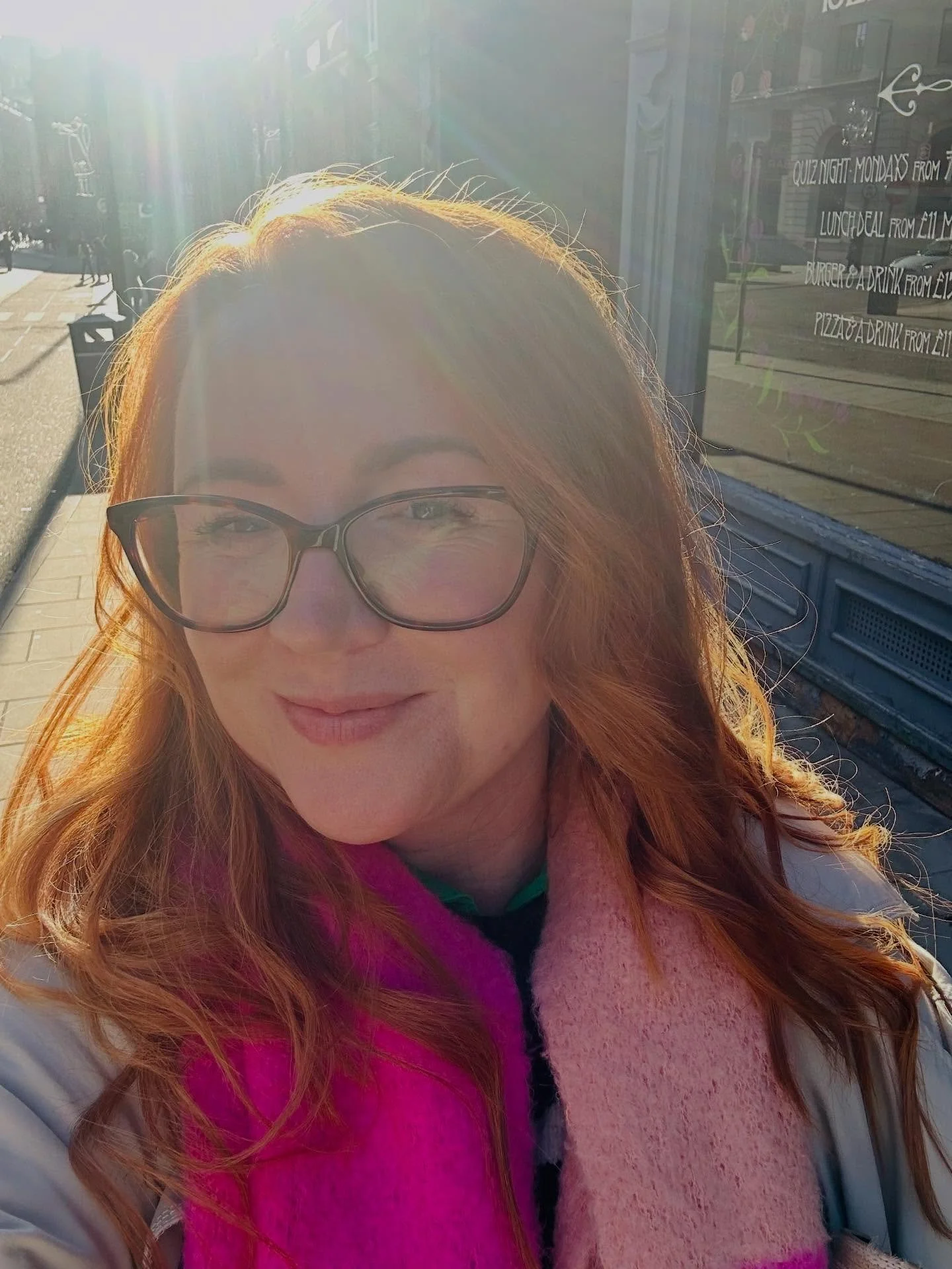 A woman with red hair, glasses, and a colorful pink and purple scarf, smiling in sunlight on a city street, in Lucy's Little Bakehouse, Dean Clough, Halifax