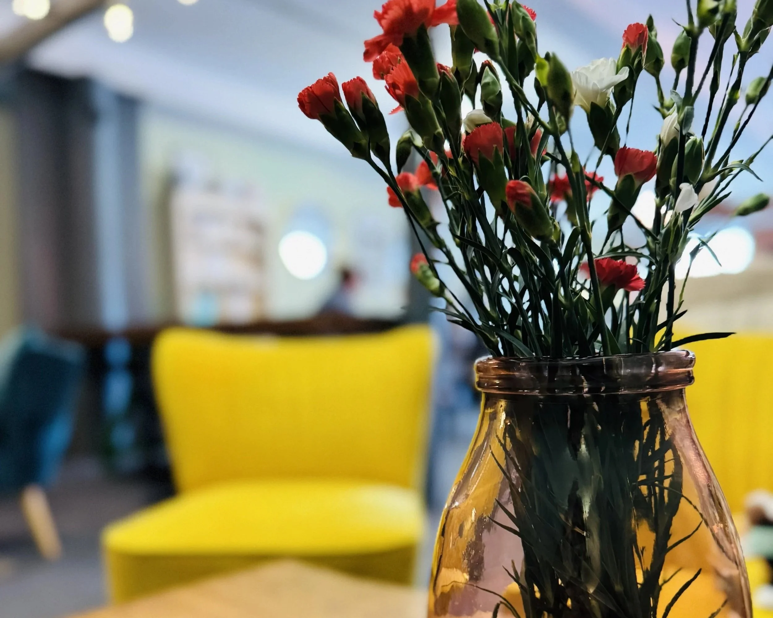 A glass vase with pink and white flowers on a table, with yellow chairs and a blurred background in Lucy's Little Bakehouse, Dean Clough, Halifax