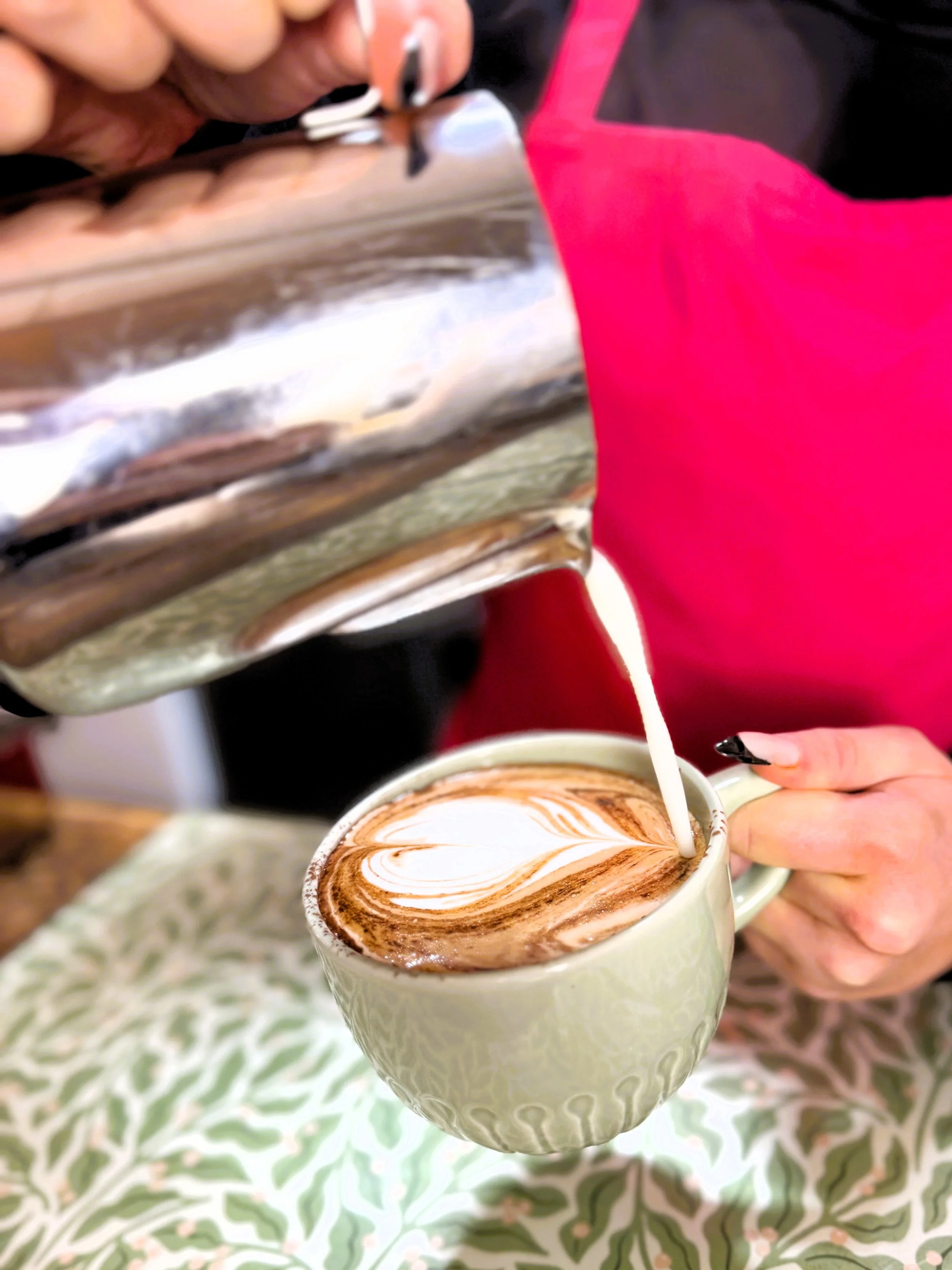 A person pouring milk into a flat white with swirled milk foam, held in their hand, on a decorative tablecloth in Lucy's Little Bakehouse, Dean Clough, Halifax.