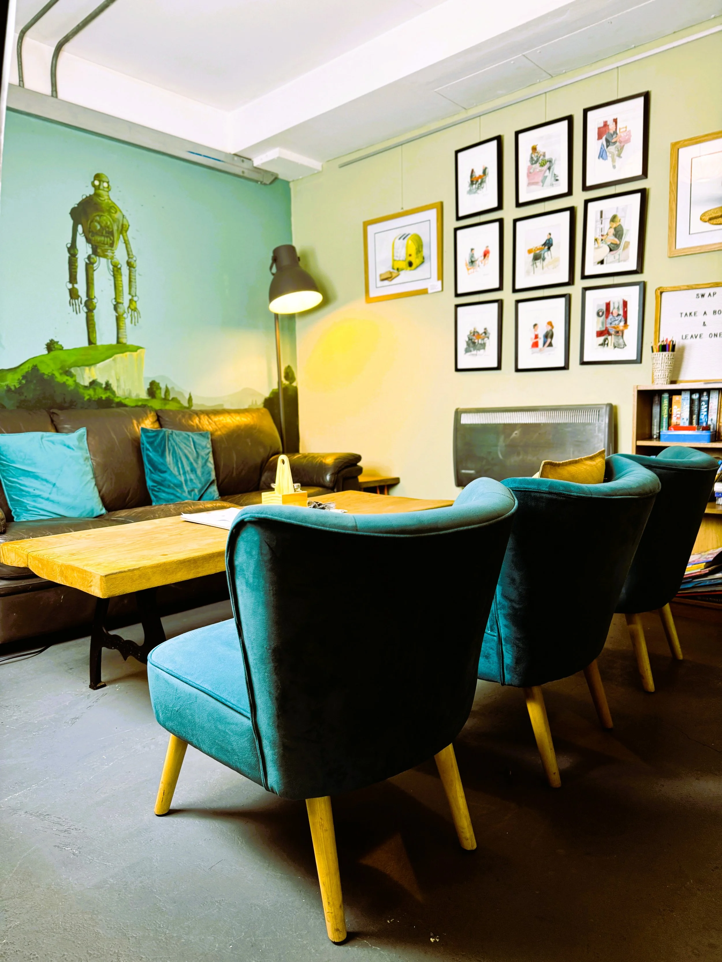 Cosy cafe with a sofa, teal pillows, matching teal velvet chairs with wooden legs, a wooden coffee table, wall art including a wall painting of a robot and a gallery of framed pictures, a floor lamp Lucy's Little Bakehouse, Dean Clough, Halifax.