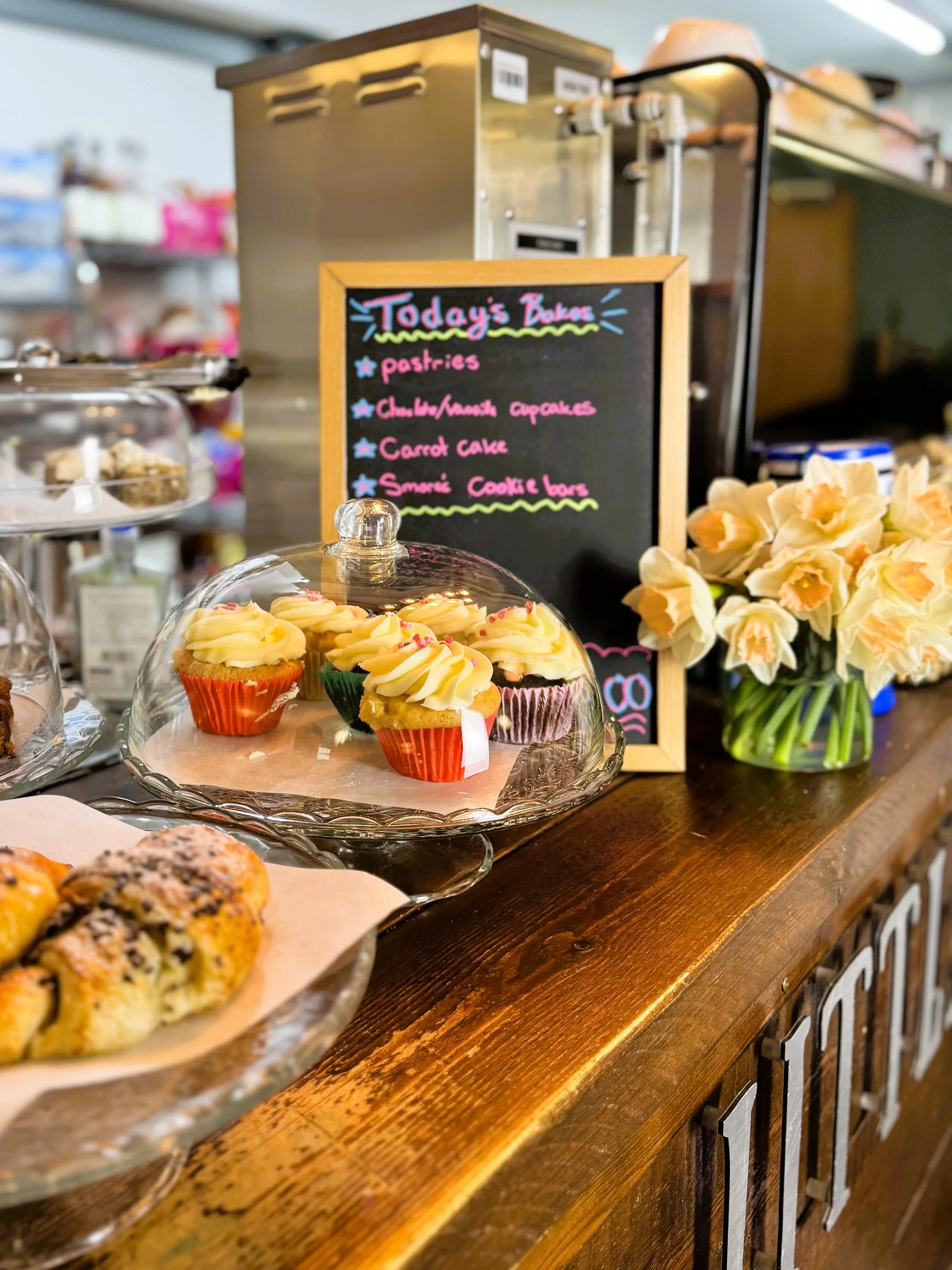 Display of cupcakes under a glass dome on a wooden counter with a chalkboard menu listing today's baked goods, flowers in a vase, and pastry treats in the background, Lucy's Little Bakehouse, Dean Clough, Halifax.