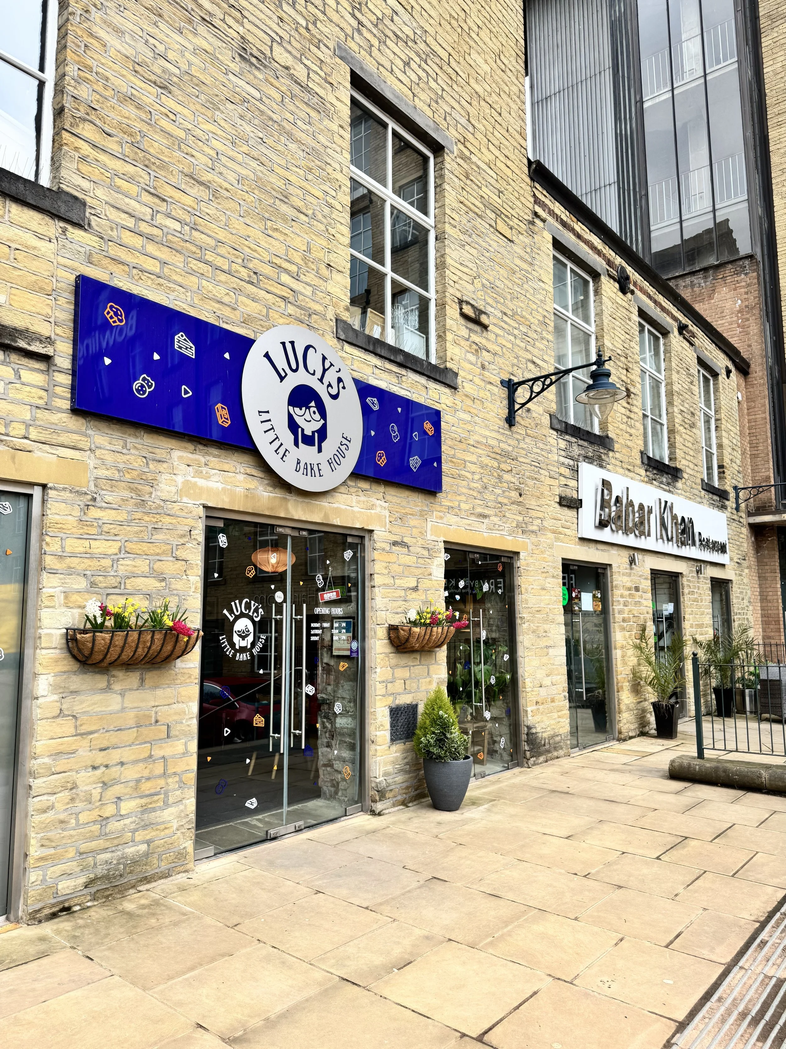 Exterior of Lucy's Little Bake House bakery storefront with glass doors, flower baskets, potted plants, and signage on a brick building. Lucy's Little Bakehouse, Dean Clough, Halifax.