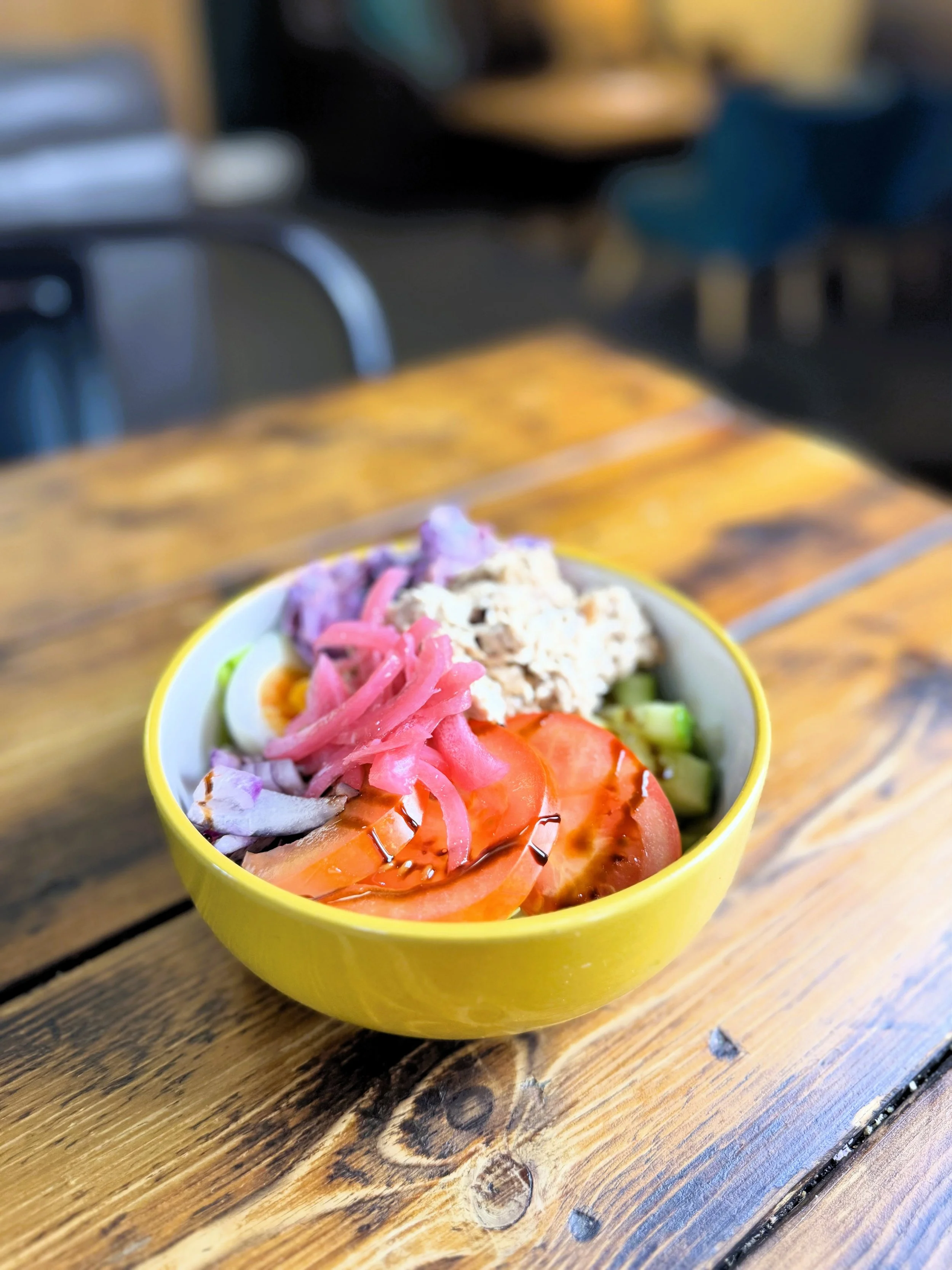 A yellow bowl containing a fresh vegetable salad with sliced tomatoes, cucumbers, pickled red onions, and a scoop of tuna salad on top, placed on a rustic wooden table Lucy's Little Bakehouse, Dean Clough, Halifax.
