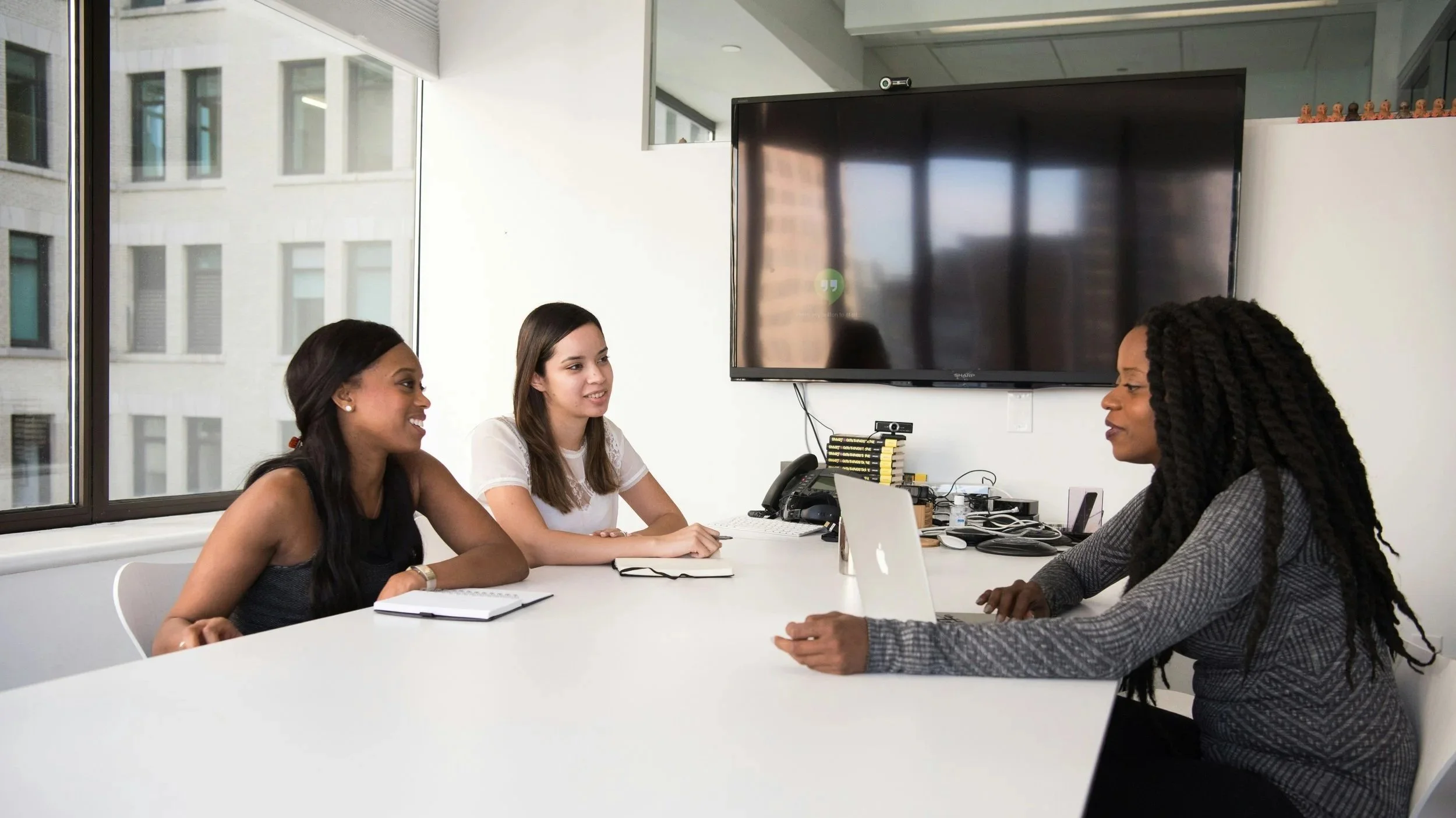 Three women sitting at a conference table, having a discussion in an office with large windows showing city buildings, and a large TV screen on the wall.