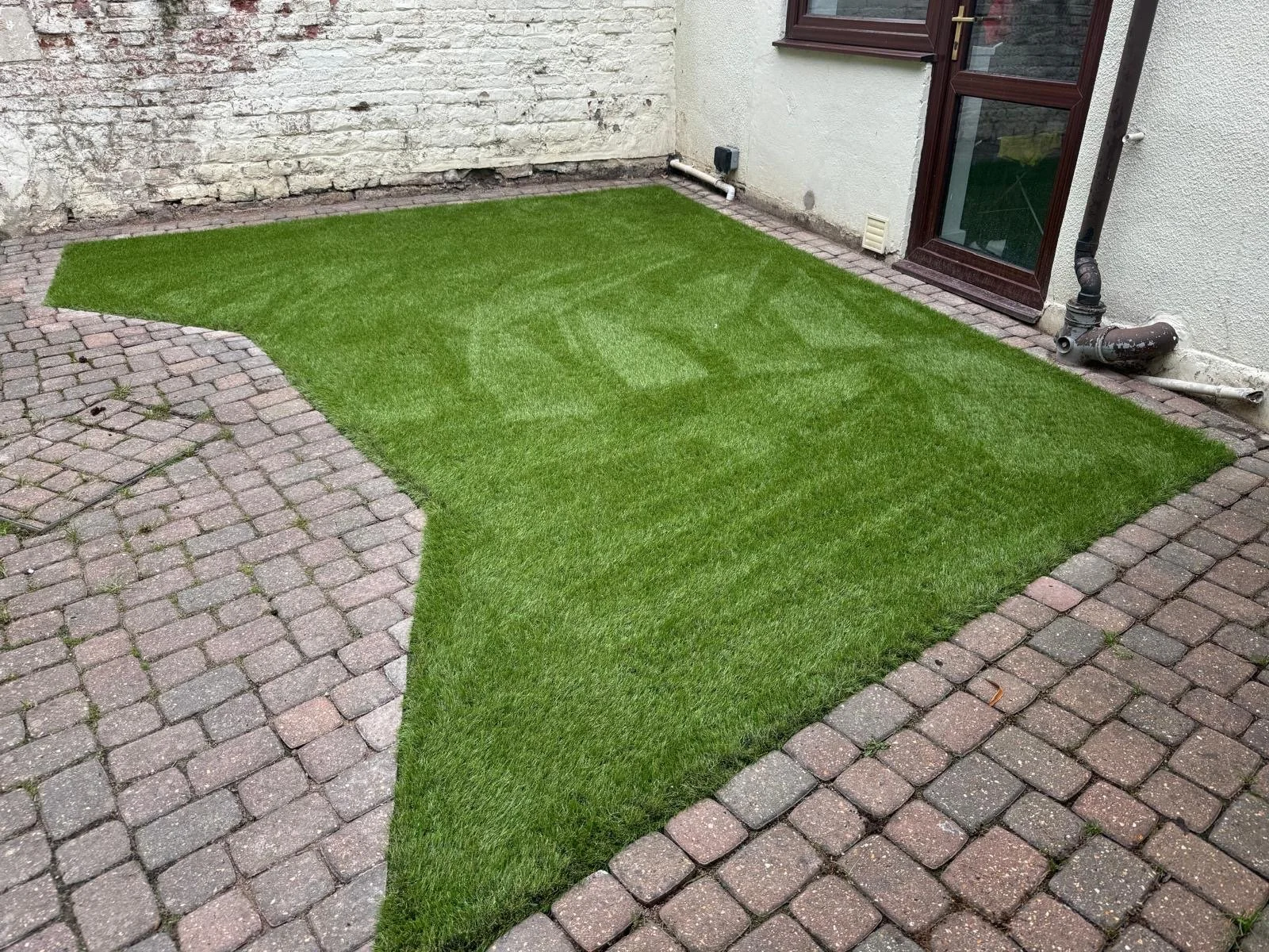 A small backyard with a patch of lush green grass surrounded by a brick-paved area and a white wall with pipes and a window.