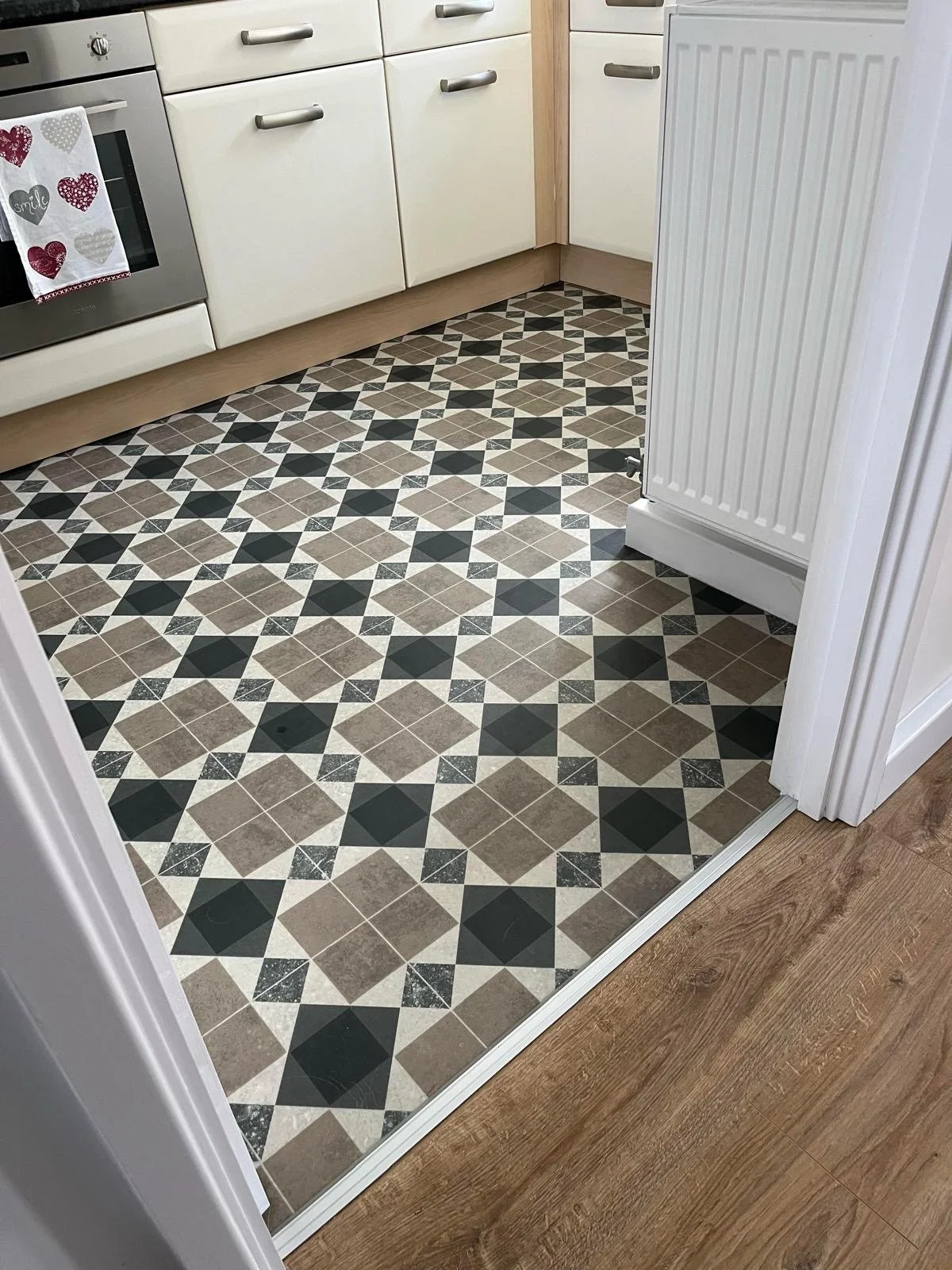Kitchen floor with patterned tile design in black, beige, and white, adjacent to a transition strip between hardwood and tiled flooring.