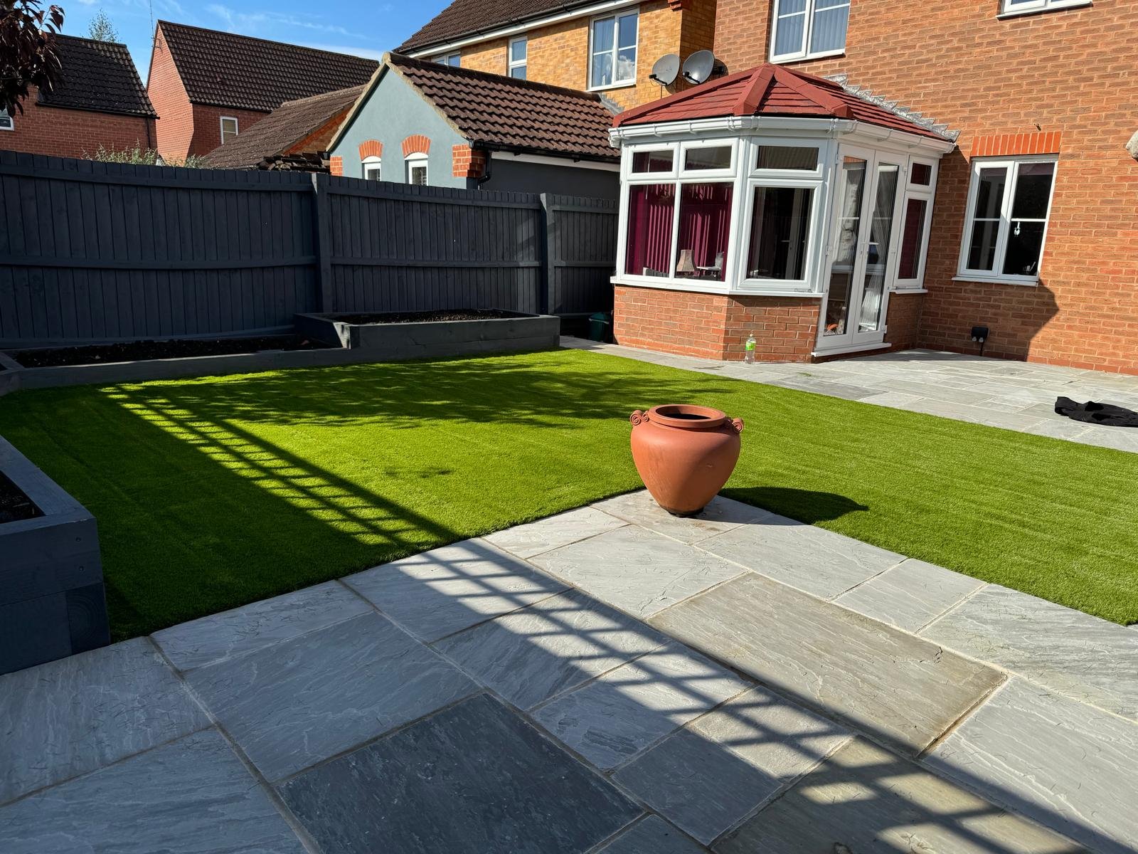 A backyard with a well-maintained artificial lawn, a paved patio, a large empty terracotta pot, a black fence, and a conservatory attached to a brick house with satellite dishes and windows.