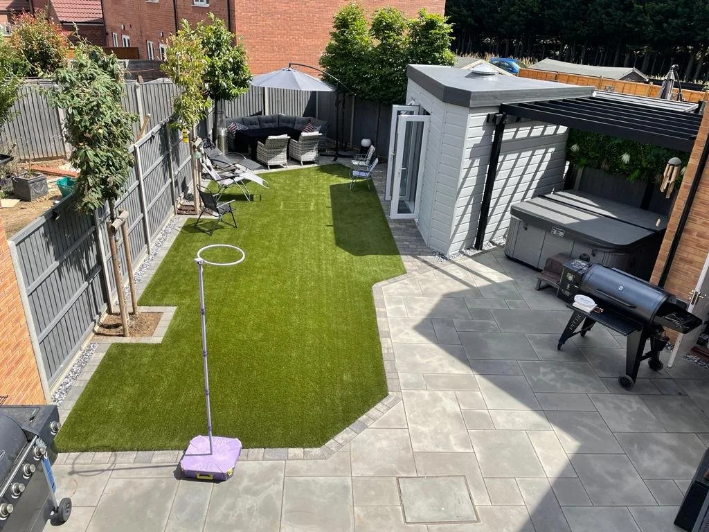 Backyard patio with artificial grass, outdoor seating, umbrella, trees, patio furniture, grill, and a white shed.