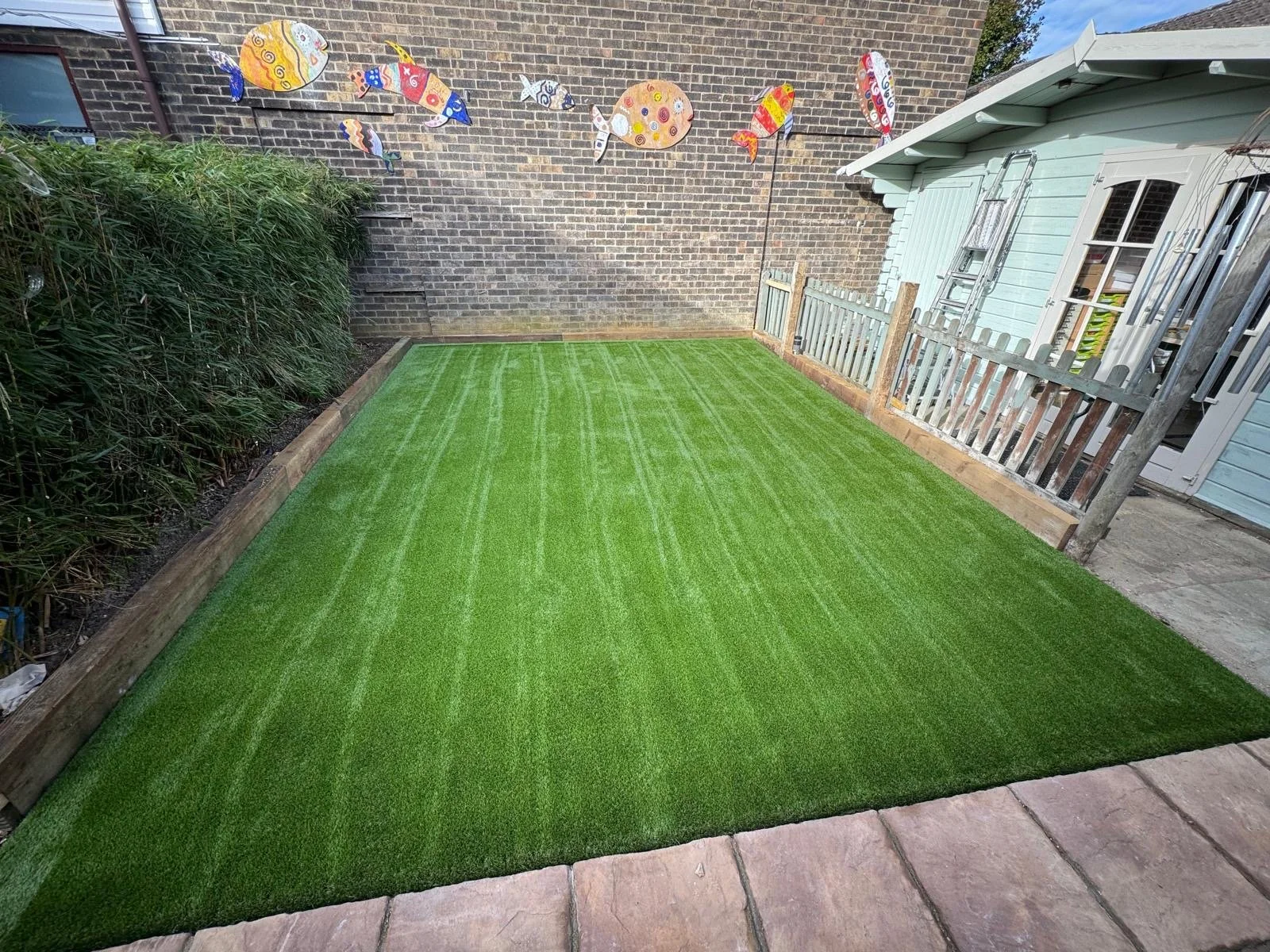 A small backyard with a freshly laid green artificial grass lawn, bordered by flowerbeds and a wooden fence on the right, a brick wall with fish-shaped decorations hanging on it in the background, and a light blue shed with a ladder against the wall.