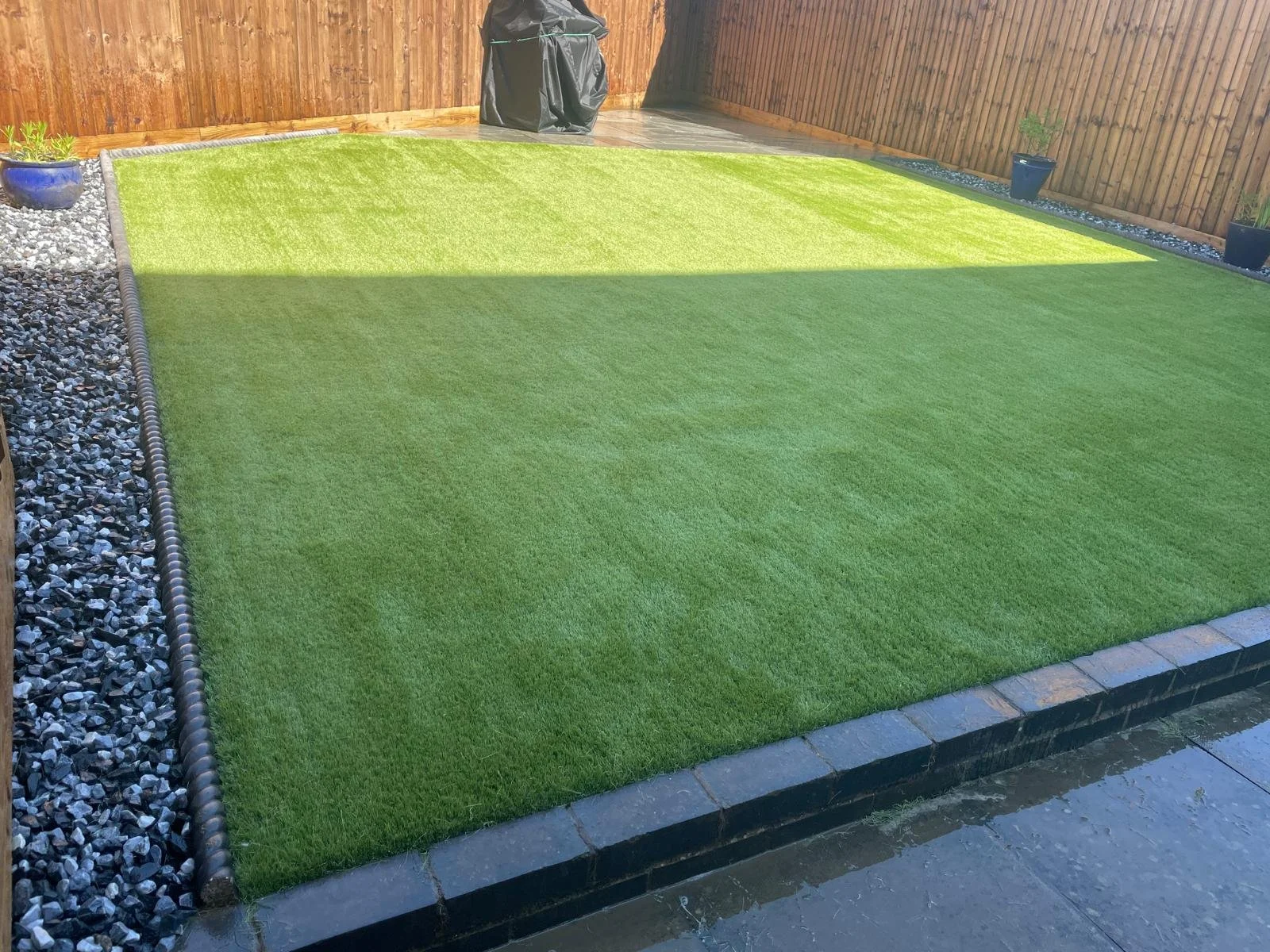 A small backyard with a well-maintained artificial grass lawn, bordered by black bricks. To the left, there is a strip of white and black decorative gravel with potted plants. In the back, there is a wooden fence, and at the corner, a covered barbecu