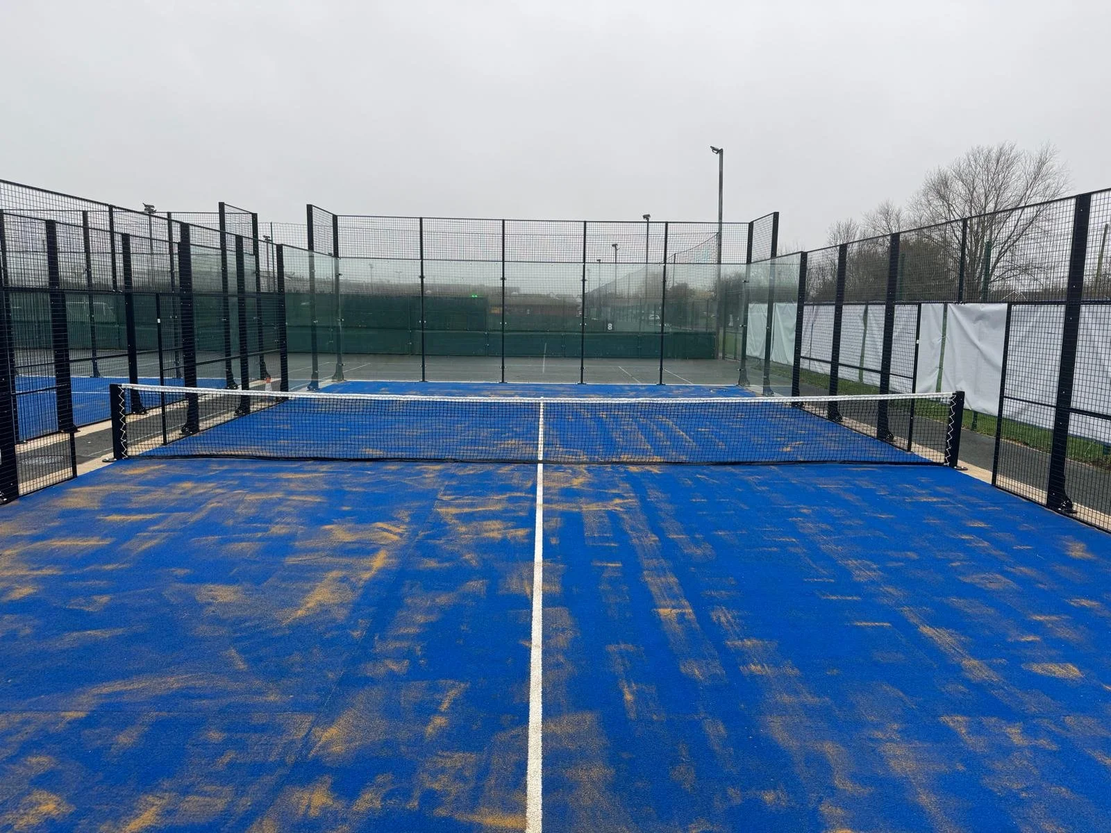 Empty outdoor pickleball court with blue surface, white lines, and black fencing on a cloudy day.