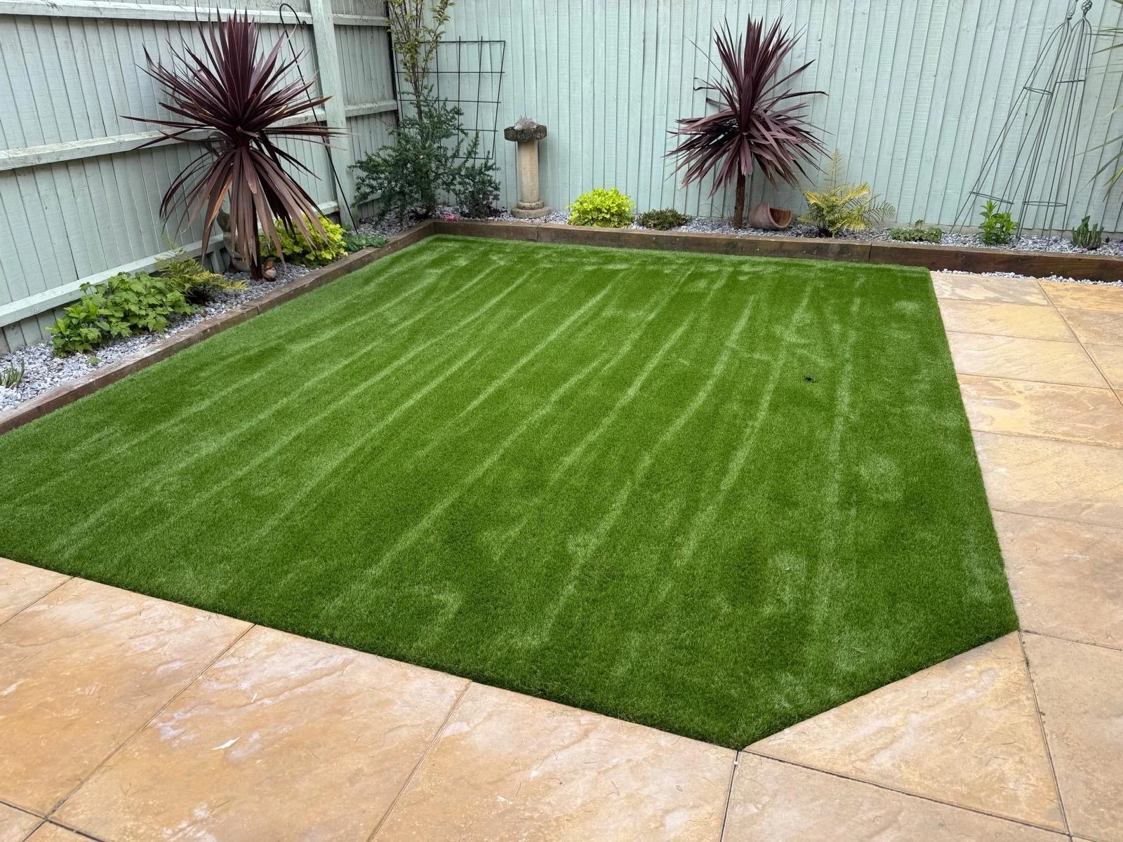 A backyard with a small green artificial grass lawn, beige tiled paving, a border of small plants, a light green wooden fence, and some decorative garden items including large purple-leafed plants, a bird bath, and a hollow ceramic pot.