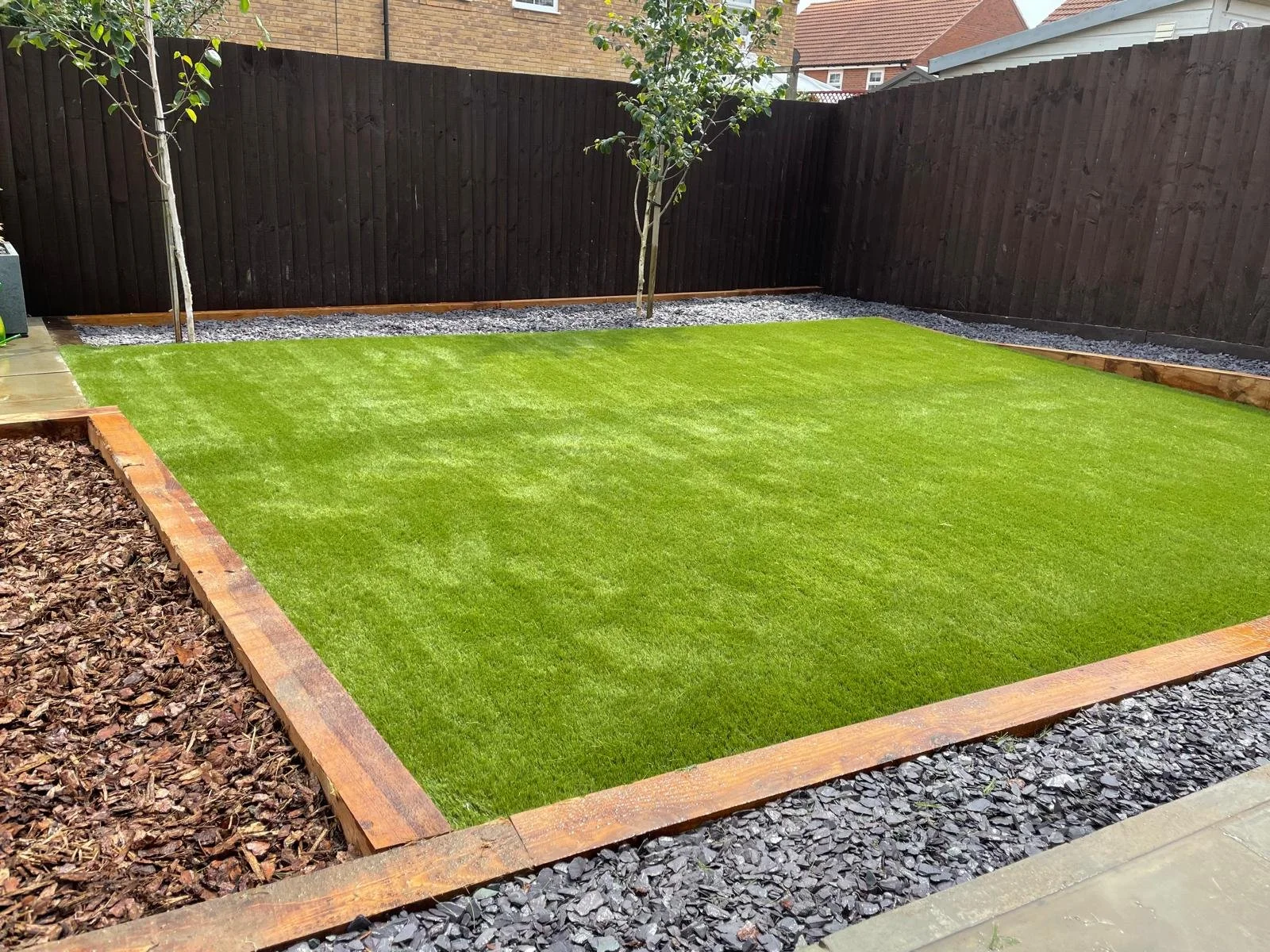 Backyard with a small lawn, two trees, a wooden fence, gravel walking paths, and existing houses in the background.