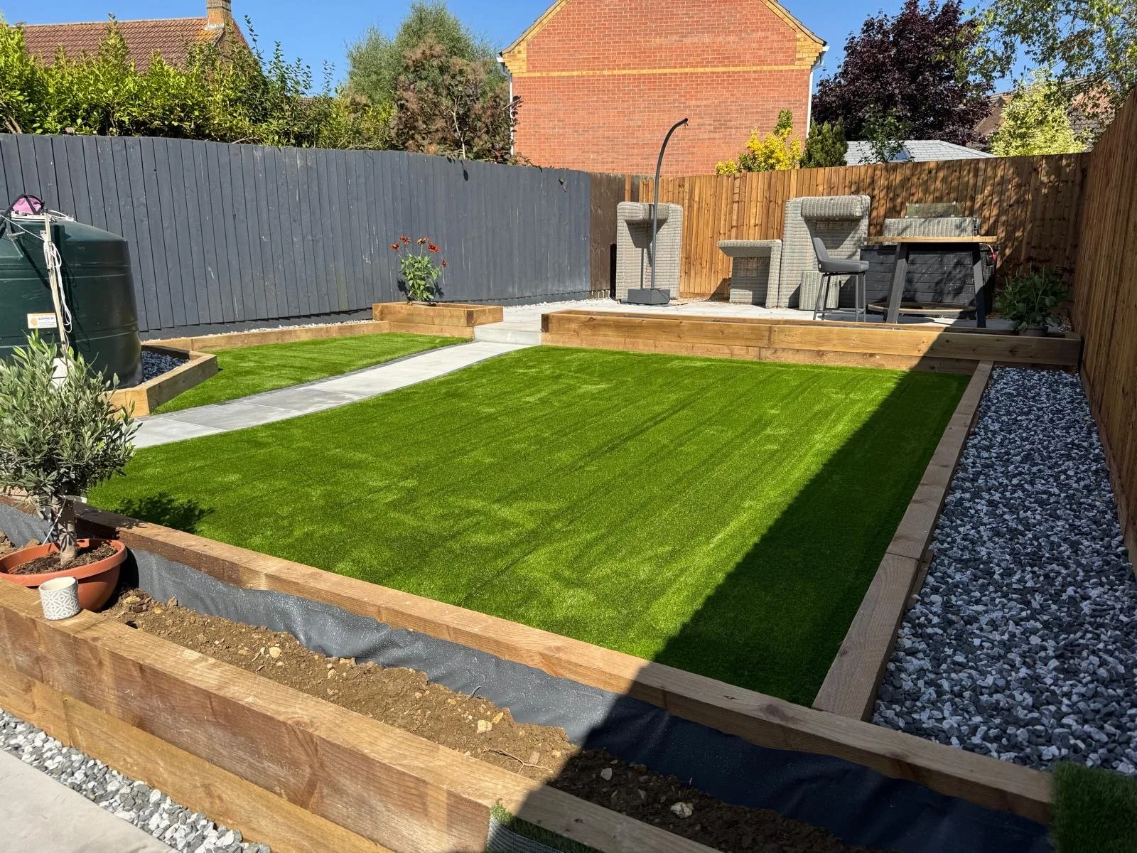 A backyard with a newly installed green artificial grass lawn, bordered by wooden edges, a stone pathway, decorative gravel, and outdoor furniture including chairs and a table on a raised wooden patio. There is a potted plant on the left and a water 