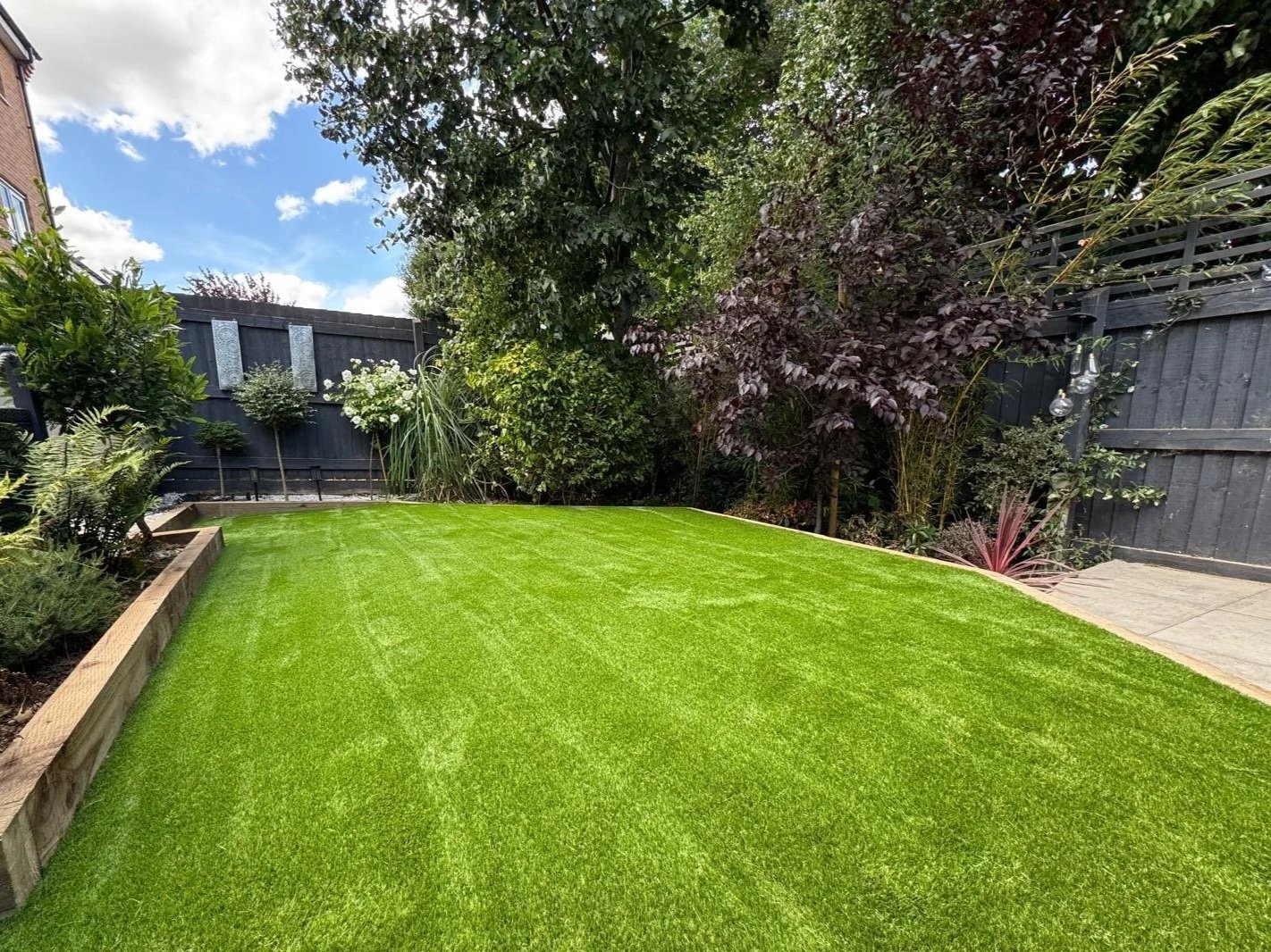 A well-maintained backyard with a lush green lawn, surrounded by various plants and trees, and a black wooden fence in the background, under a partly cloudy sky.