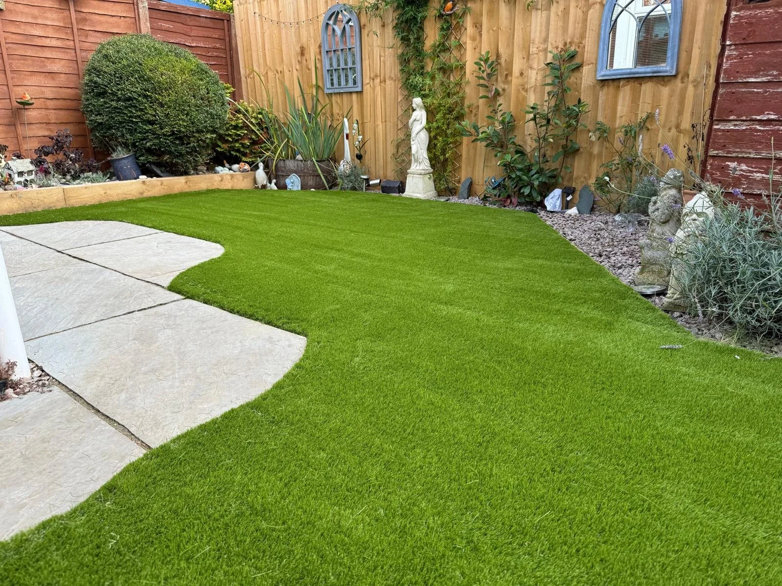 A back yard with a green artificial grass lawn, a stone pathway on the left side, and a wooden fence with decorative elements including a statue, rocks, and plants.