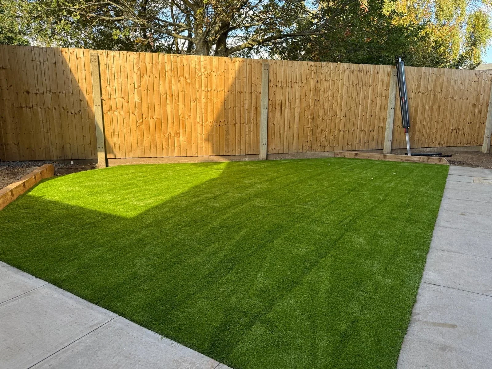 A backyard with a freshly laid green artificial grass lawn, bordered by a wooden fence and a concrete walkway on the right side. There is a tree behind the fence and a construction tool leaning against it.