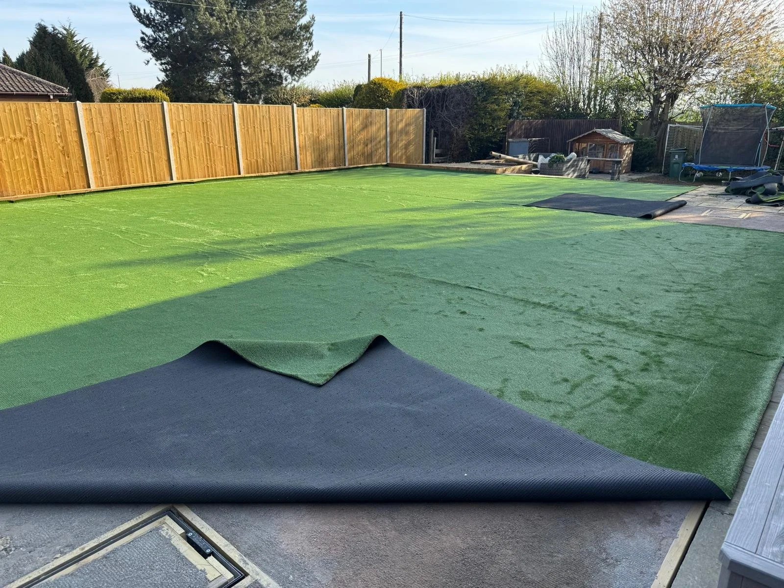Backyard with a newly laid artificial grass lawn, partially rolled out, enclosed by a wooden fence, and with various outdoor items and equipment visible.
