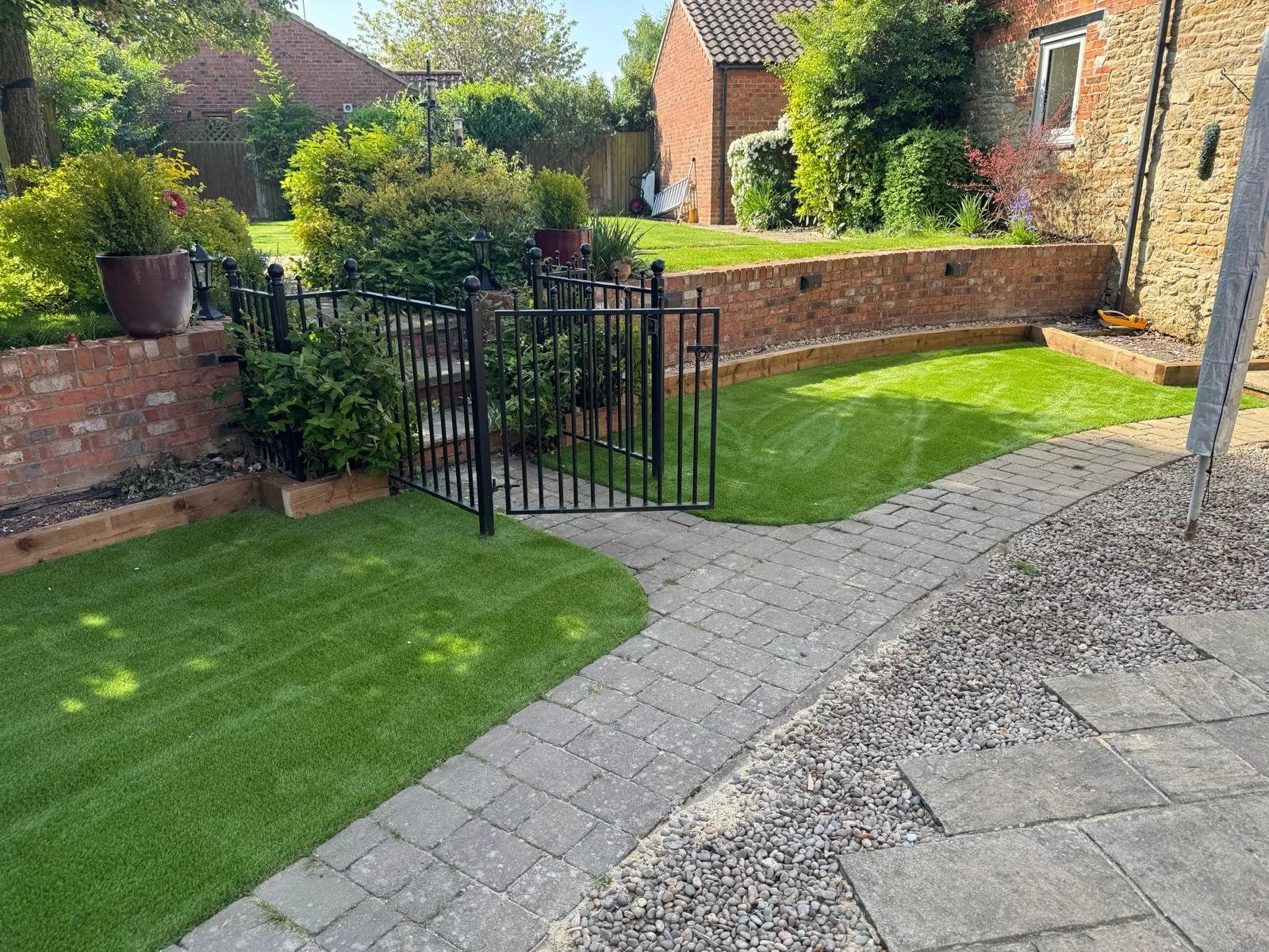 A backyard garden with a maintained green lawn, a curved brick wall, a small gate, and a paved patio area with gravel and flagstone. There are plants, shrubs, and potted flowers along the wall, and a neighboring brick house in the background.