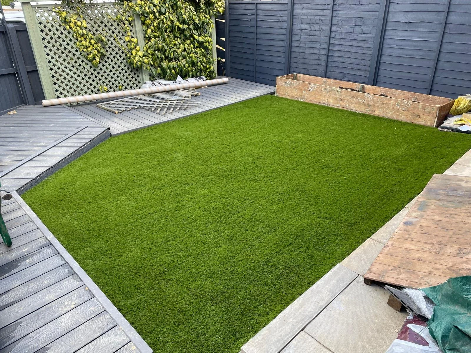 A backyard with newly installed green artificial grass, surrounded by wooden decking and fencing, with some gardening tools and supplies visible.