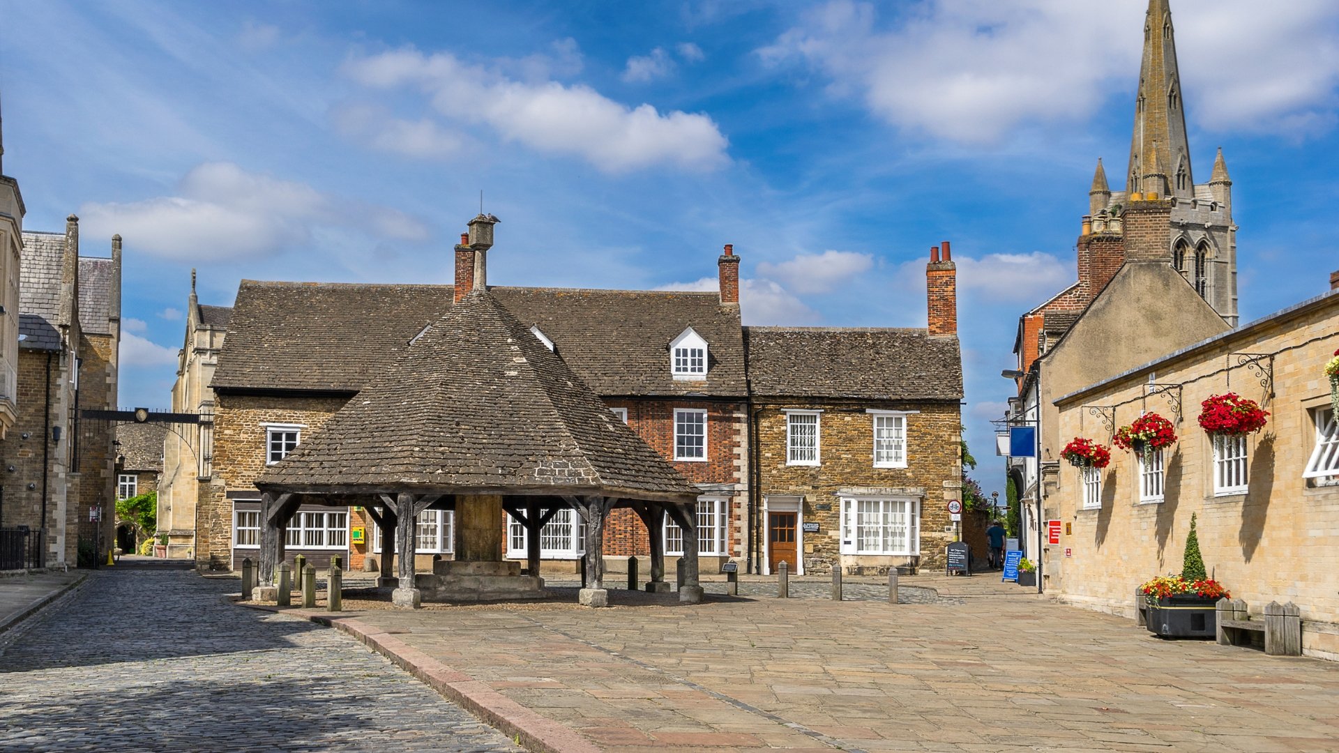A historical town square with cobblestone streets, old stone buildings, a well in the center, and a church tower in the background under a partly cloudy sky. Red flowers are hanging from the building on the right.