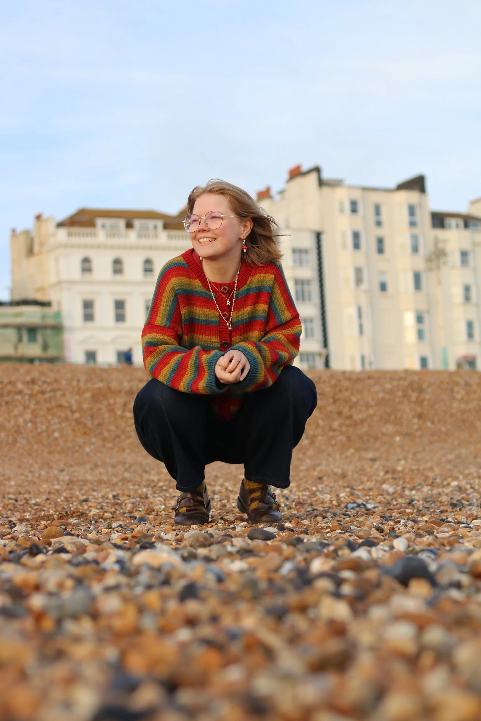 A young woman with shoulder-length blonde hair and glasses, crouching on a pebbled beach in front of a row of white buildings, smiling and wearing a colorful rainbow striped sweater.