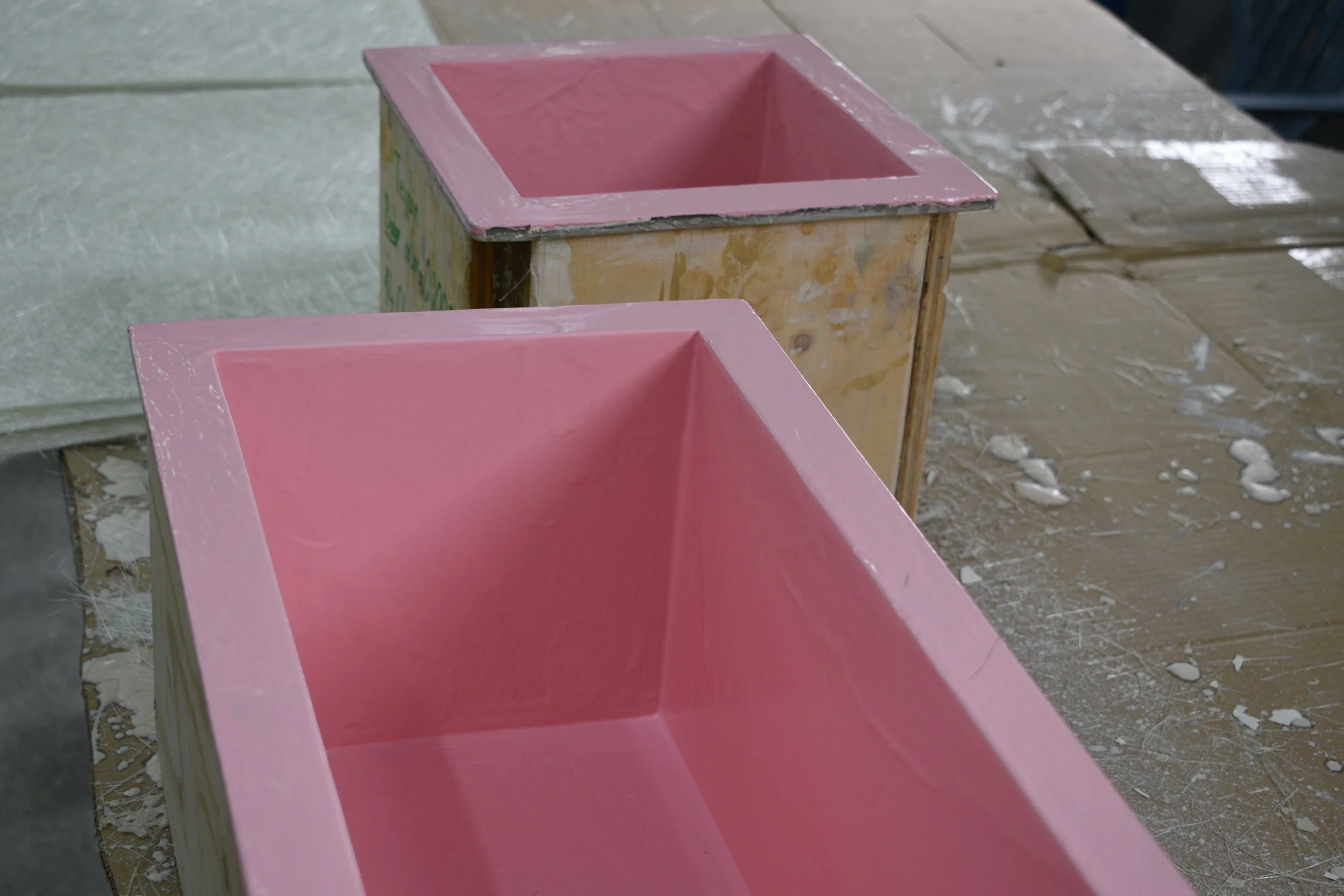 A close-up of two wooden rectangular-shaped molds painted pink, with a workshop or craft room background, tools, and scattered materials.